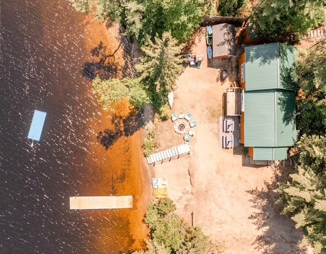 Aerial view of a lakeside property with dock, boat, trees, and a building with a green roof.