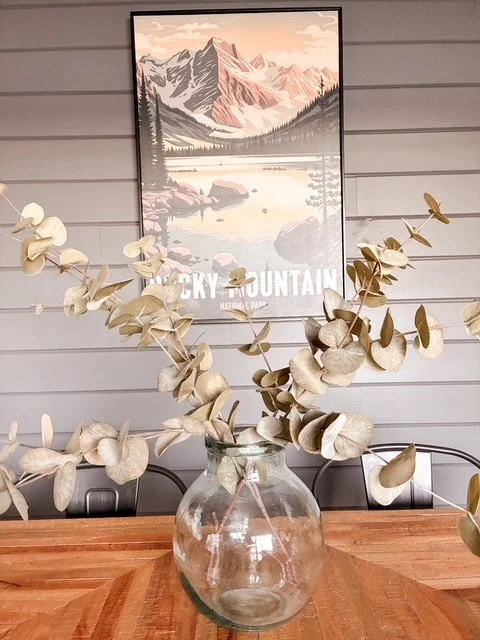 Vase with dried eucalyptus branches on a wooden table, with a mountain landscape poster on a gray wall in the background.