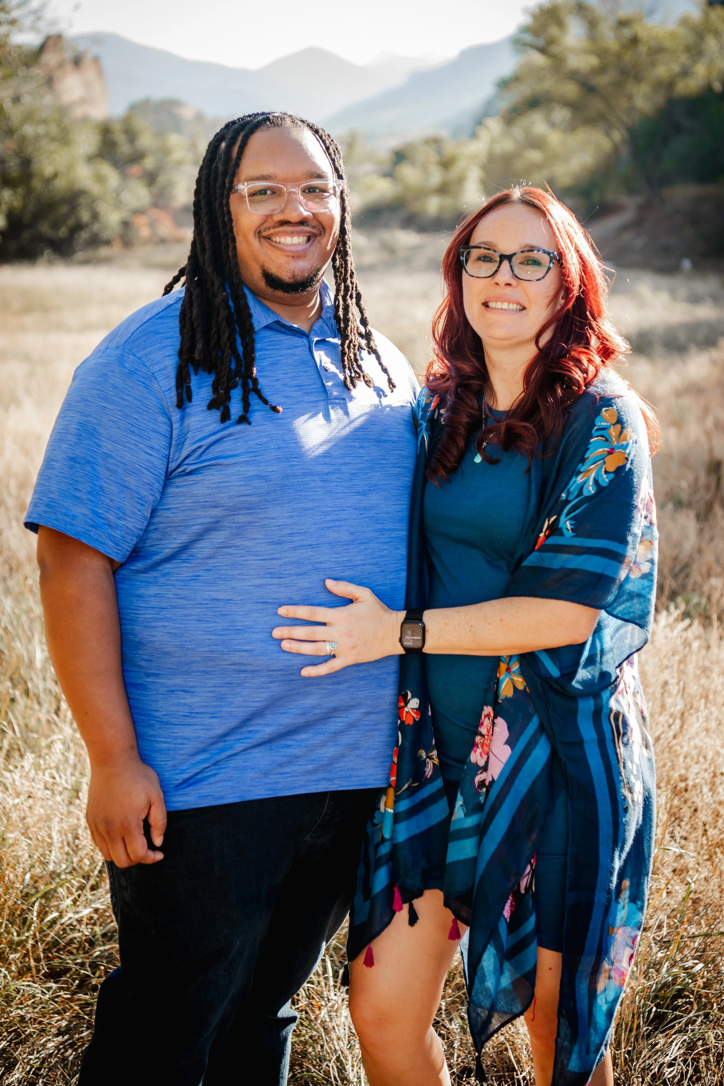 A smiling couple standing outdoors in a field with mountains and trees in the background, dressed in blue clothing.