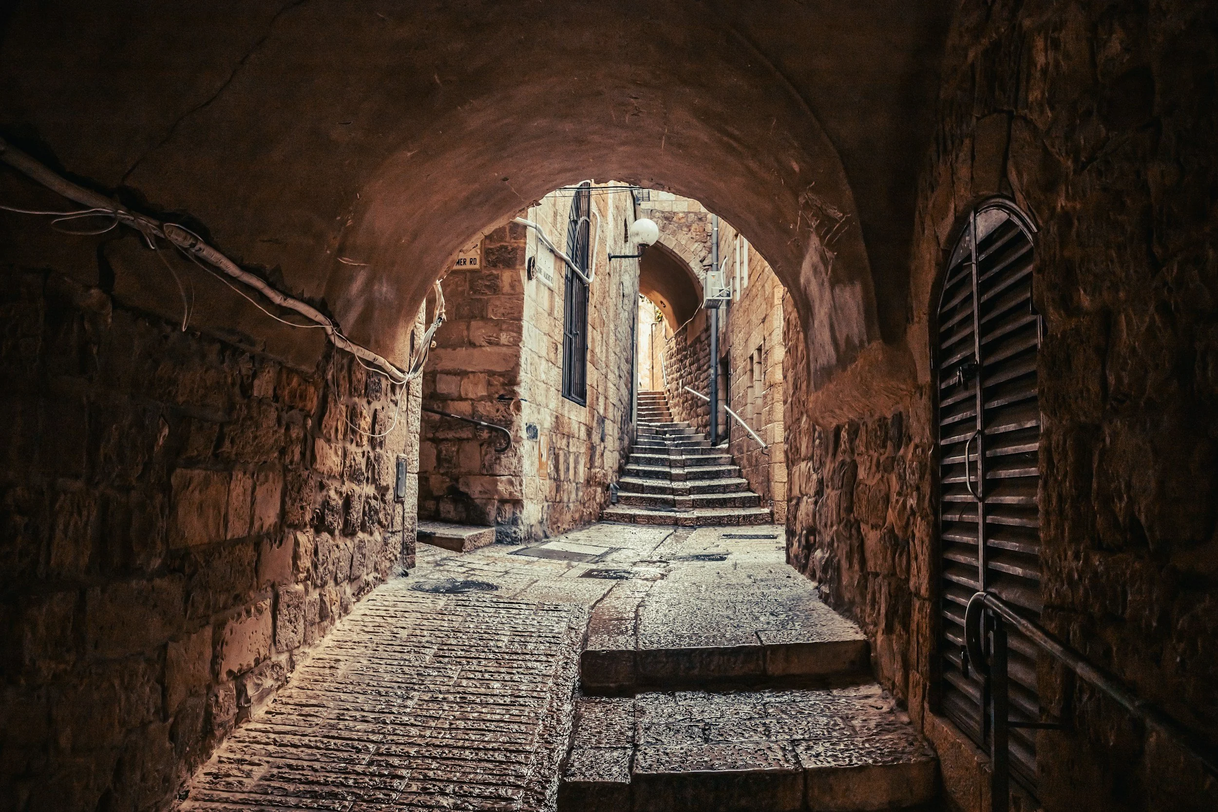 Stone alleyway in the old city of Jerusalem with archway, stairs, and brick walls, illuminated by sunlight.