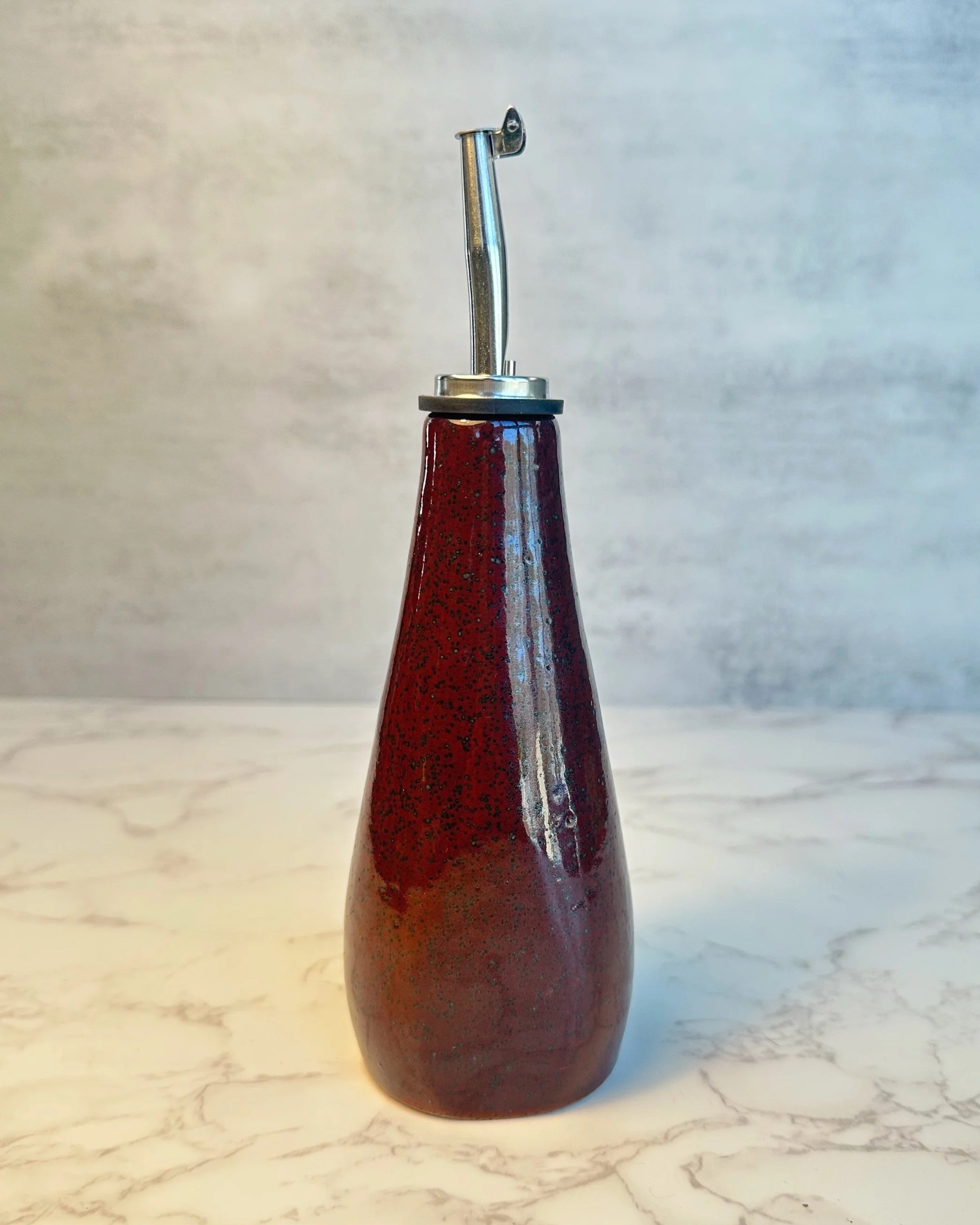 Wheel-thrown ceramic oil bottle with stainless steel pour spout in deep red stoneware glaze