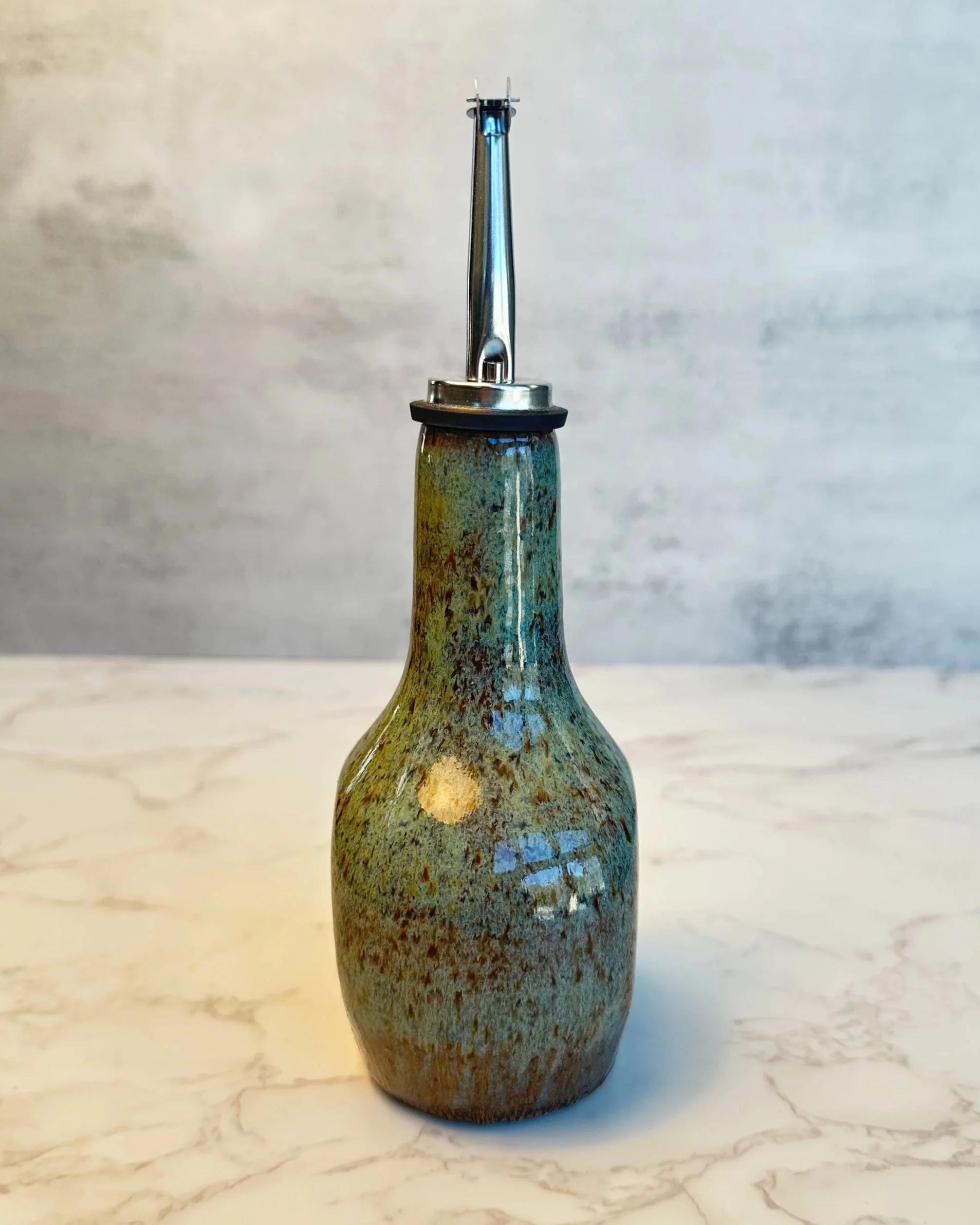 Side view of handmade ceramic oil dispenser with stainless steel pour spout in earthy green tones and speckled clay