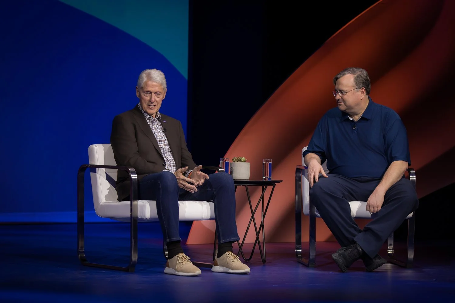 Two men sitting on white chairs on a stage, engaging in conversation. One is wearing a dark blazer, checkered shirt, jeans, and beige sneakers, while the other is dressed in a blue polo shirt and dark pants. There is a small black table with a potted