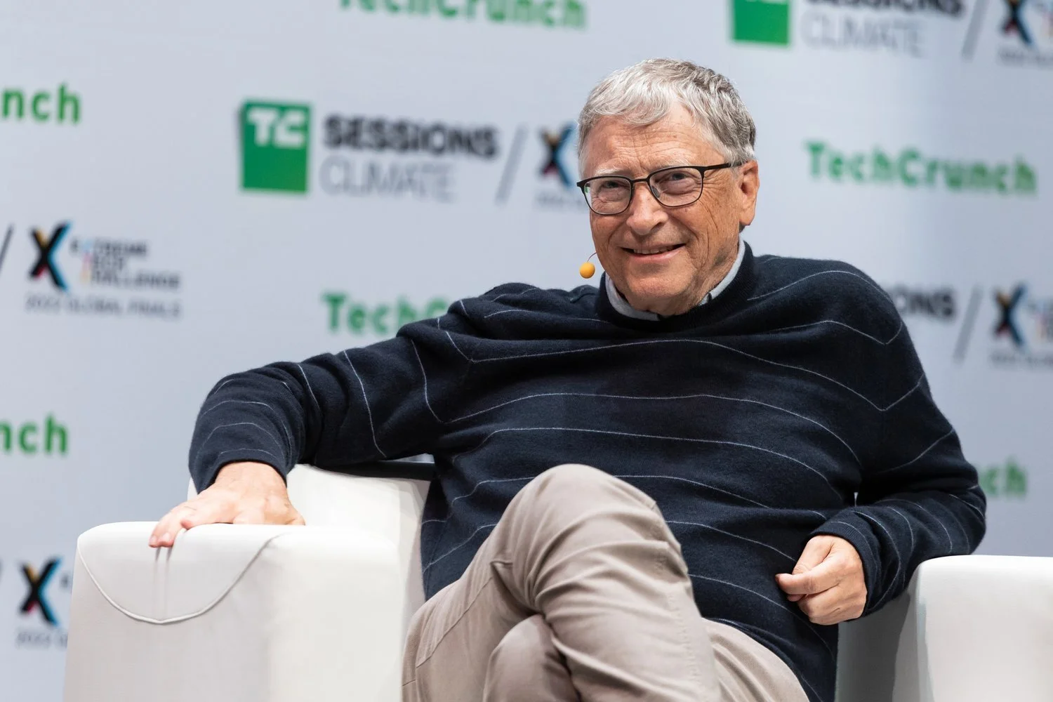 Bill Gates sitting on a white chair, smiling, wearing glasses, a dark sweater with white stripes, and beige pants, during a conference with a backdrop displaying the TechCrunch logo and other event branding.