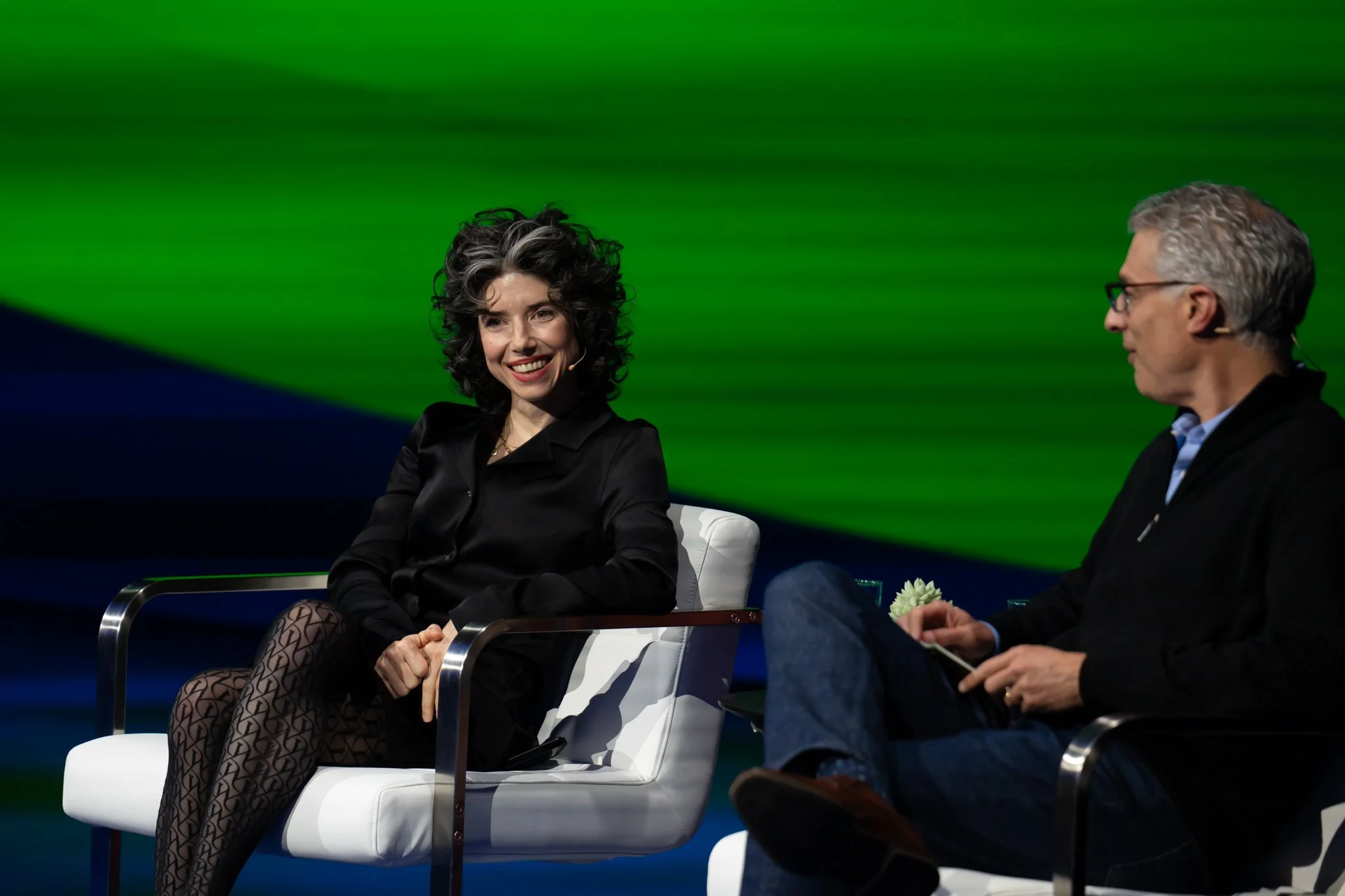 Two people sitting and talking on stage, with a green and blue background. The woman on the left has curly dark hair and is smiling, wearing a black blouse and patterned stockings. The man on the right has short gray hair, glasses, and is wearing a d