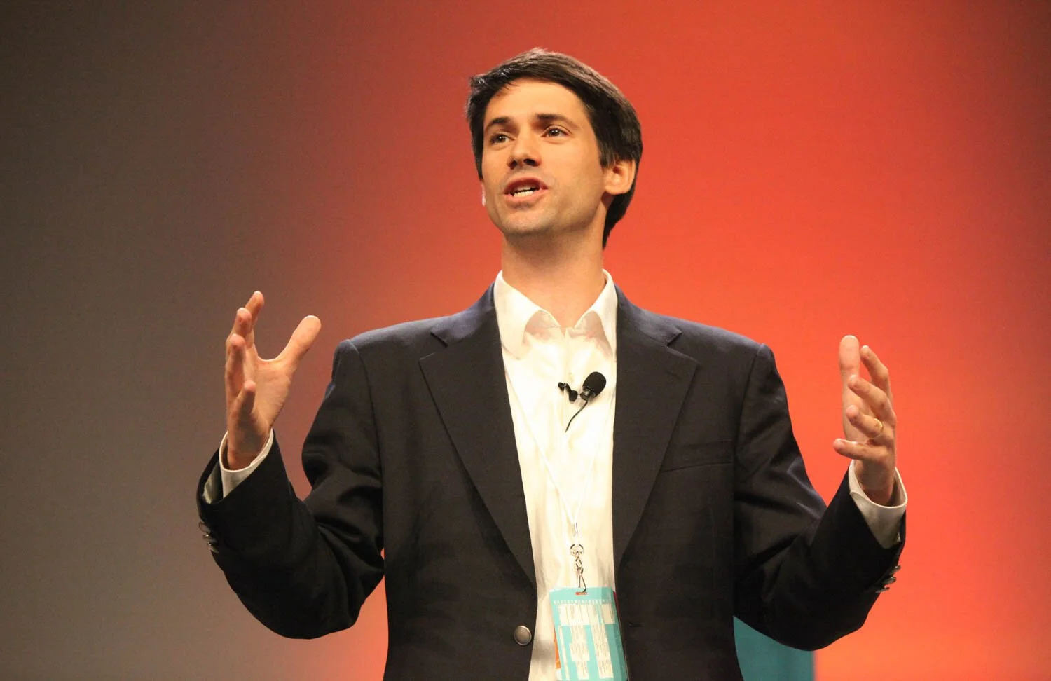 Man in a black suit and white shirt giving a presentation on stage with a red and black background.
