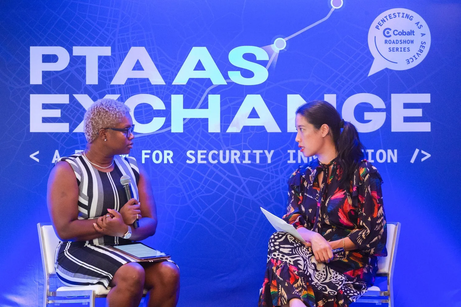 Two women sitting on stage talking at a PTAAAS exchange event focused on security information, with a blue backdrop displaying the event's name and a map design.