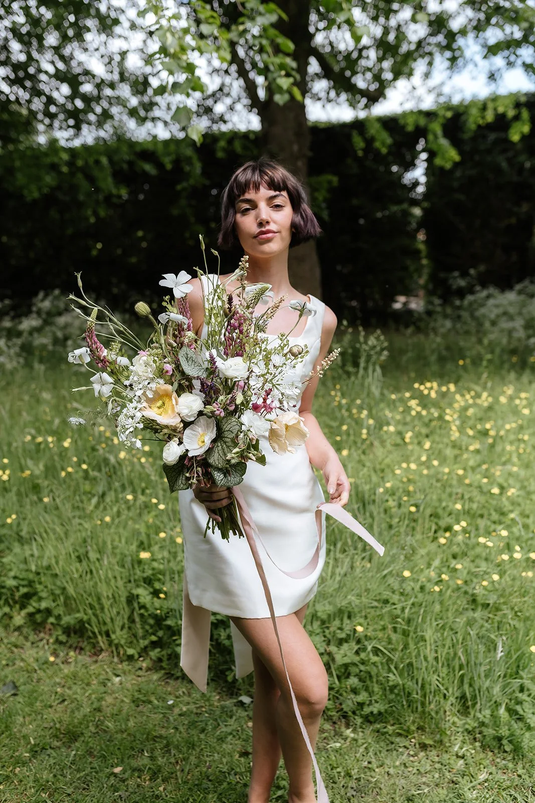 A young woman in a white dress holding a large bouquet of flowers outdoors on a grassy area with trees in the background.