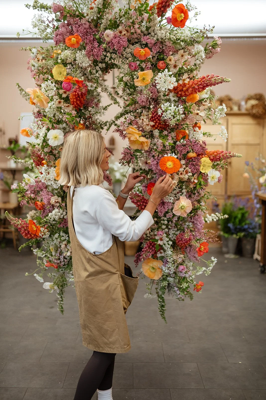 A woman with blonde hair arranging a large floral arch with pink, orange, white, and yellow flowers inside a flower shop.