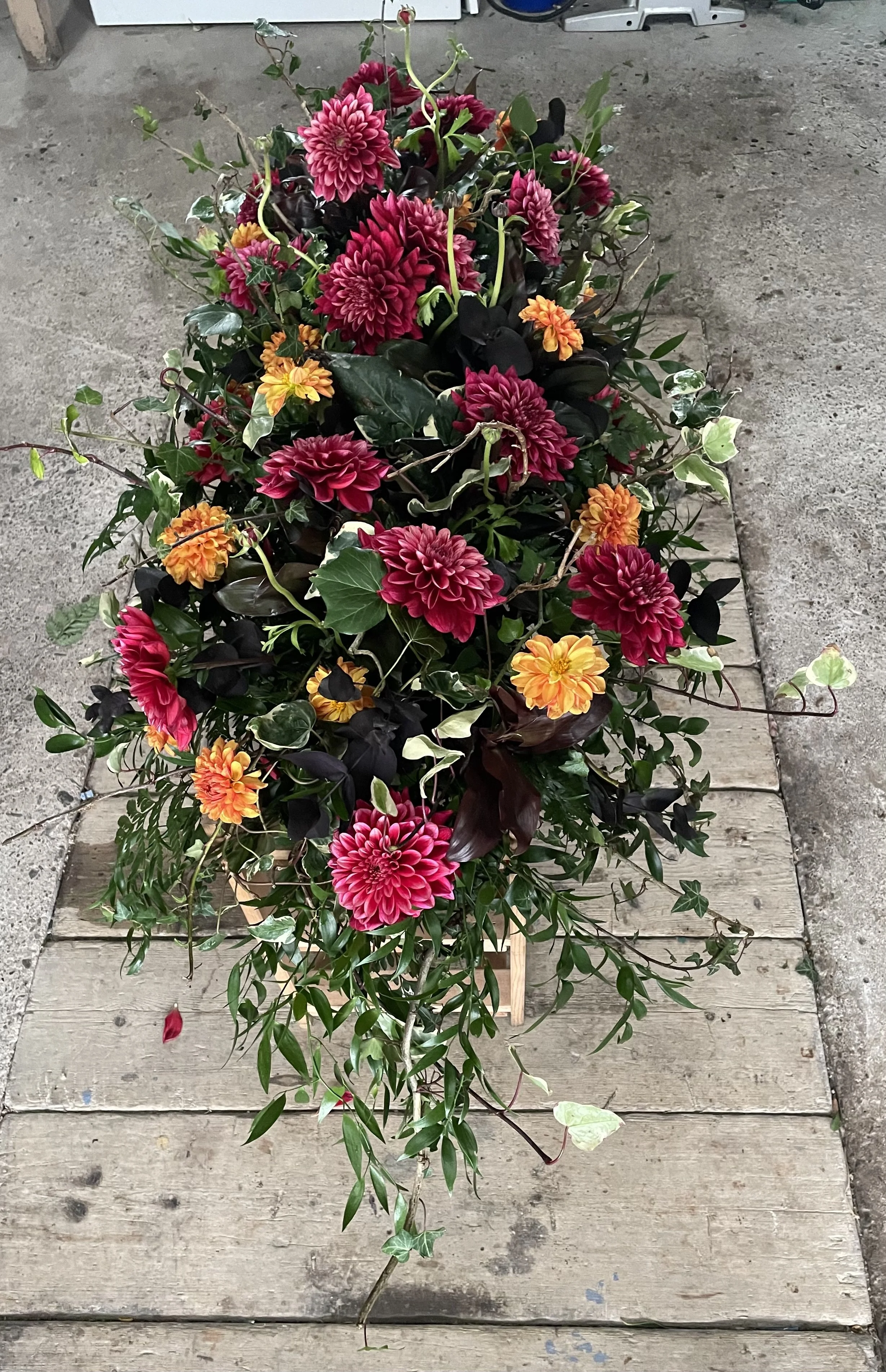A floral arrangement of pink dahlias, orange marigolds, and green foliage on a wooden plank on a concrete surface.