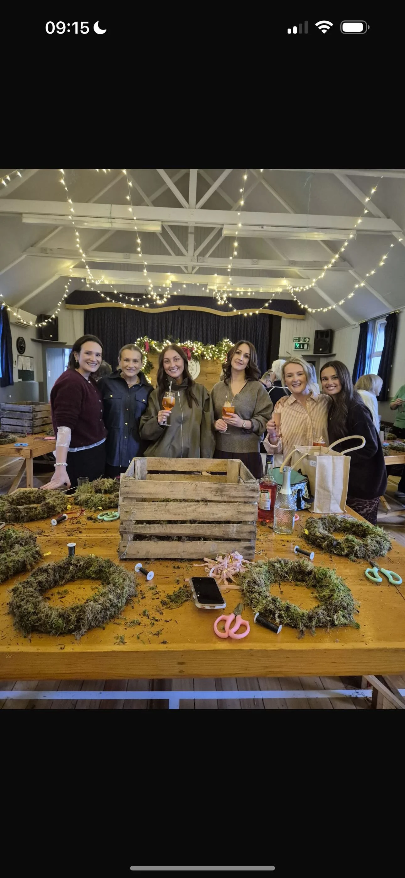 Six women standing behind a table with wreaths, crafting supplies, and drinks during a holiday celebration with Christmas lights and decorations in the background.