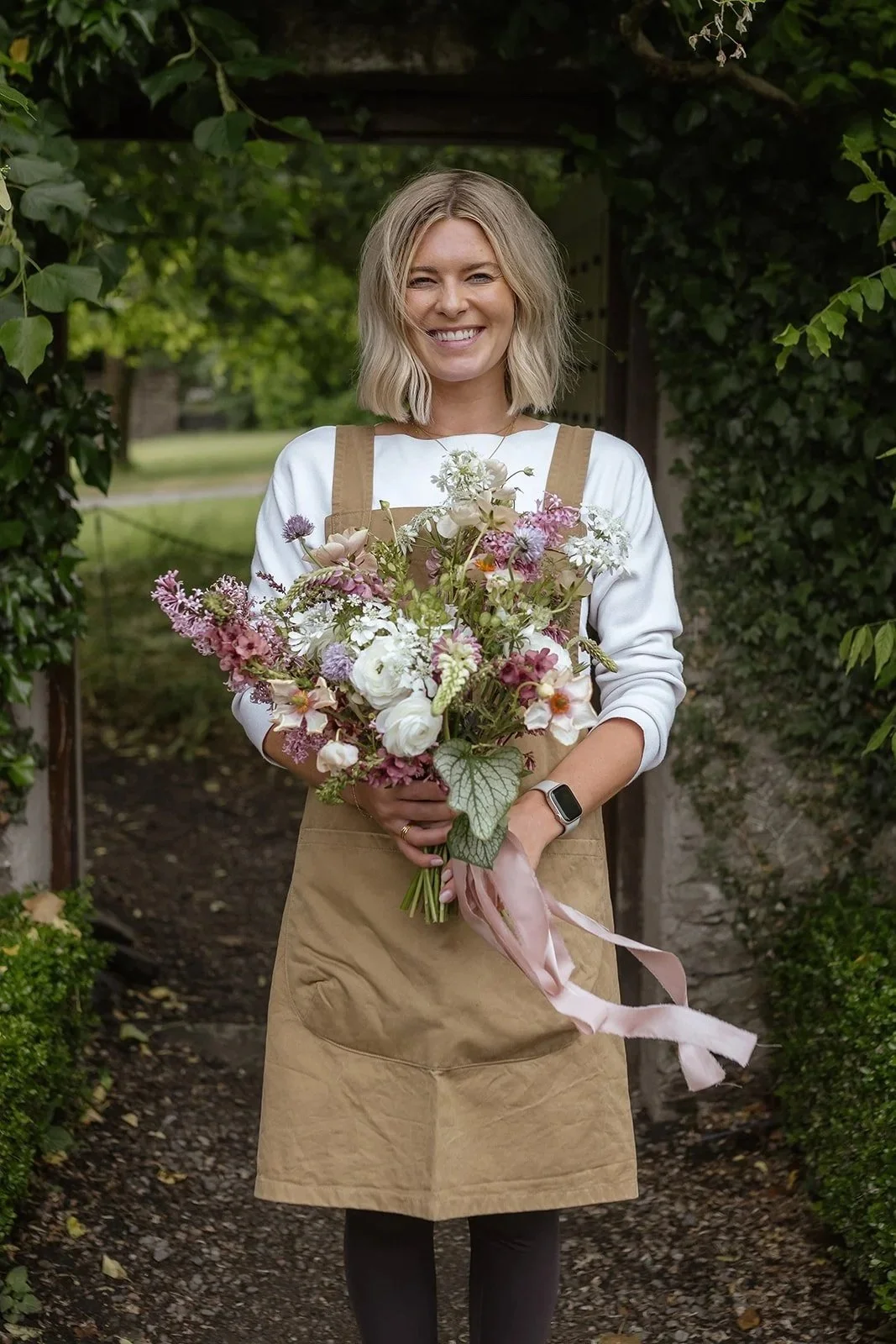 A woman with blonde hair and a smile, holding a bouquet of pink, white, and purple flowers, standing outdoors in a green garden setting.