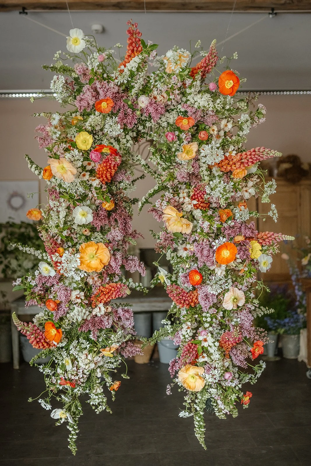 A large floral arch decorated with pink, orange, yellow, white, and purple flowers, set indoors against a ceiling and a wooden cabinet in the background.