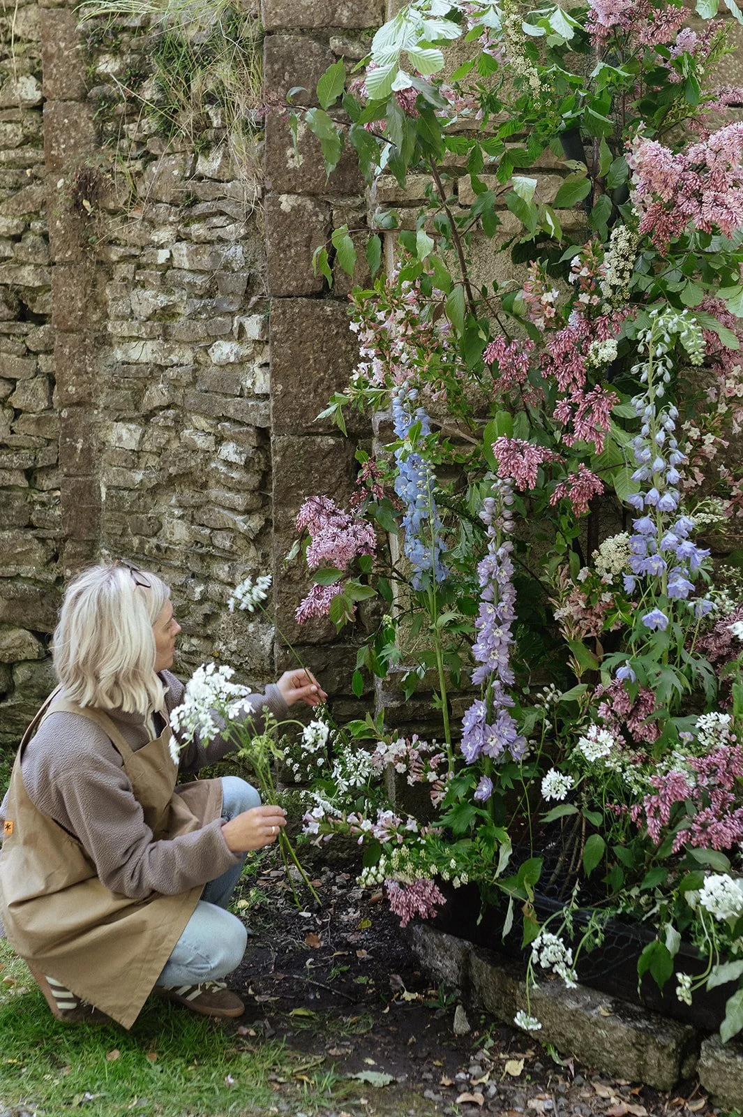 A woman with blonde hair kneeling on the grass, holding flowers, looks at a large display of colorful flowers against a stone wall.