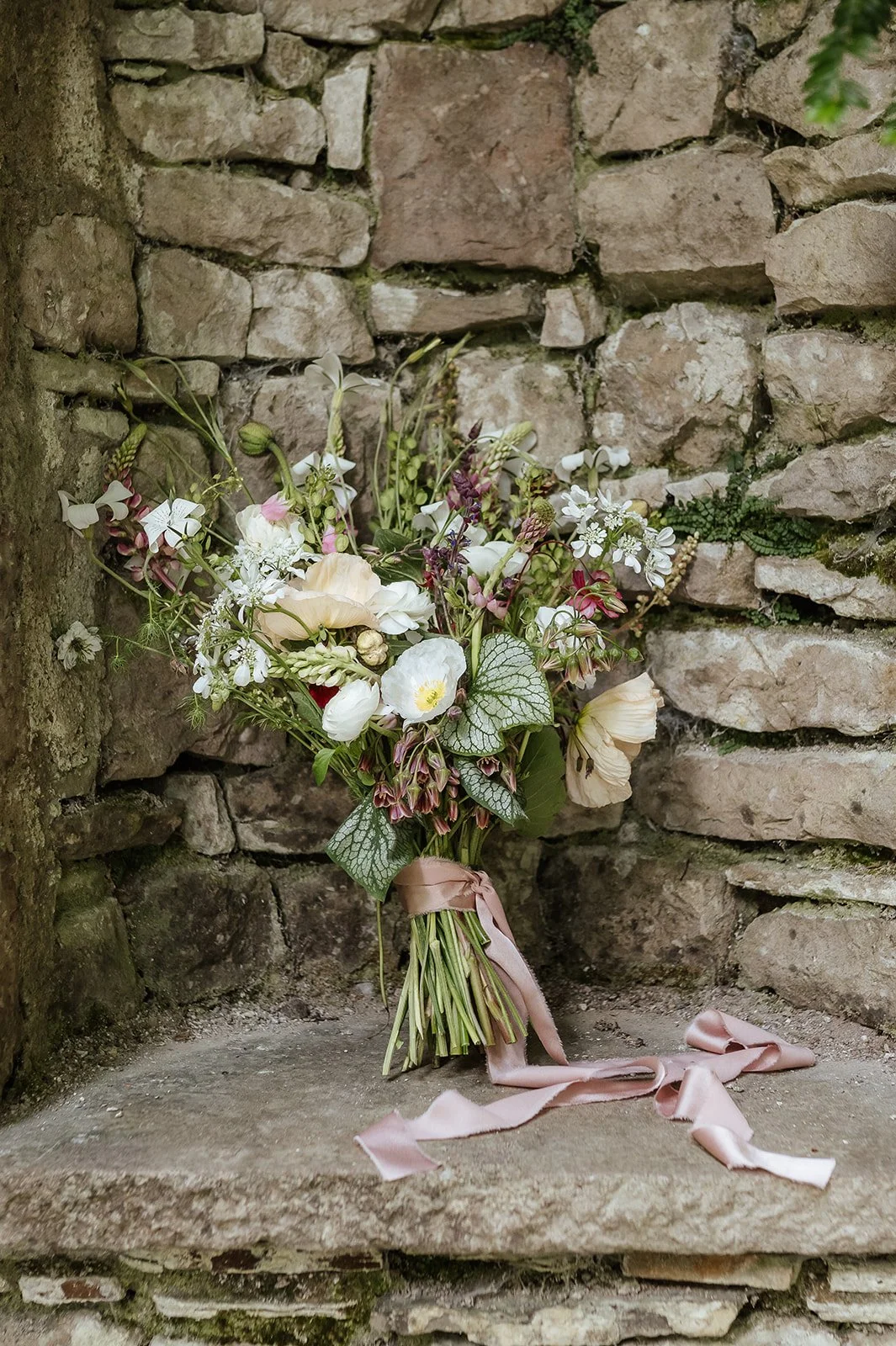 A bouquet of mixed flowers including white, cream, and pink blooms with green leaves, tied with a pink ribbon, resting on a stone ledge against a rustic stone wall.
