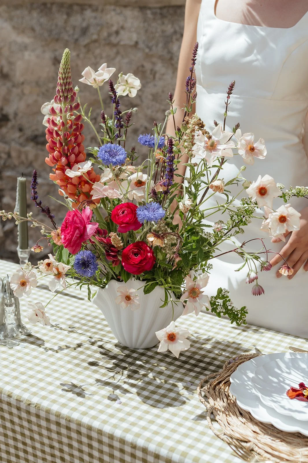 A white vase filled with colorful mixed flowers on a checkered tablecloth, with a person in a white dress standing nearby.