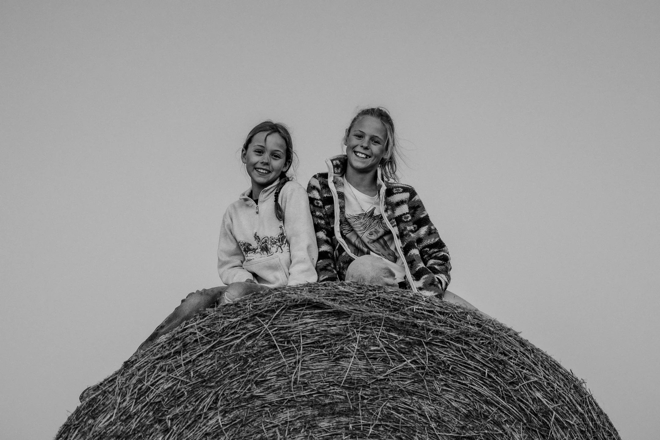 two young girls sit on top of a round bale of hay, smiling