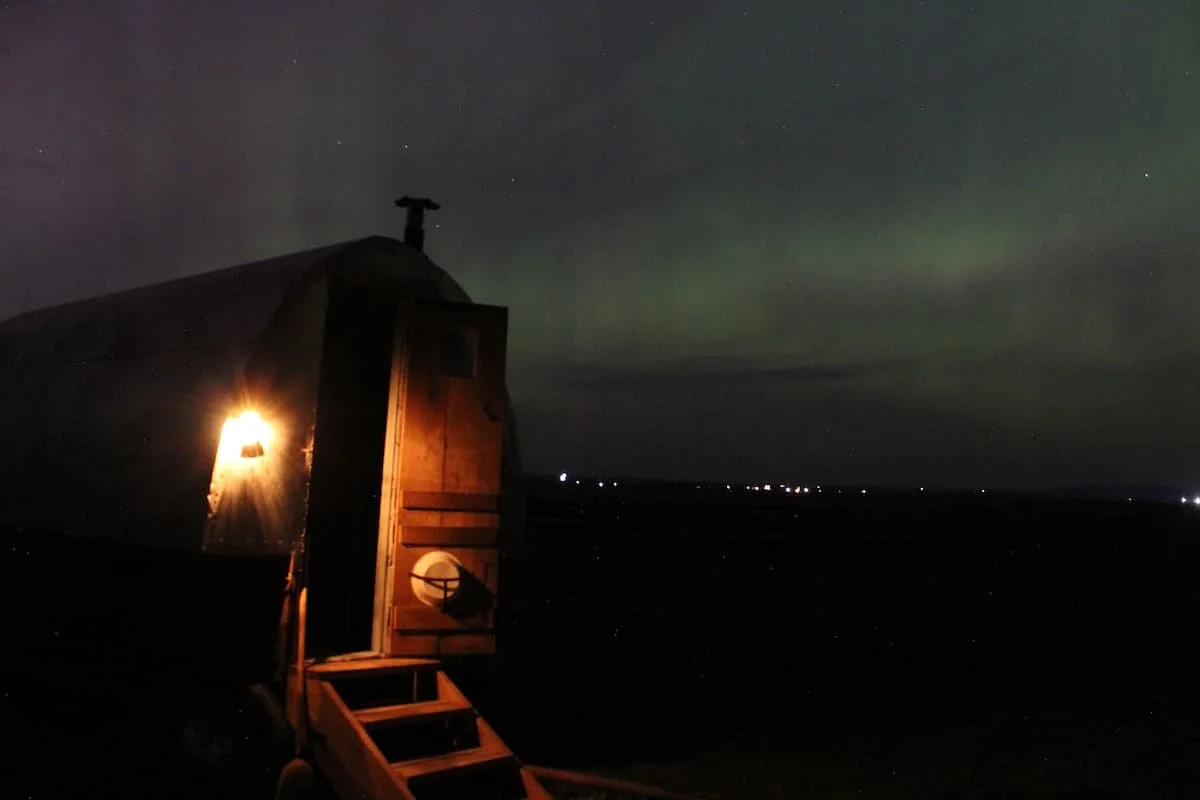 A lantern glows in the dark on the front of a vintage sheepwagon while the northern lights display as green and purple in the background