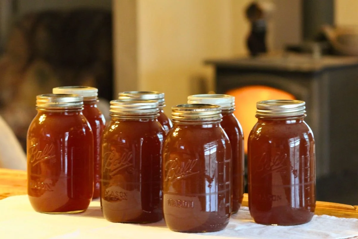 Seven jars of chicken bone broth sit on a white towel with a fire burning in a wood stove in the background