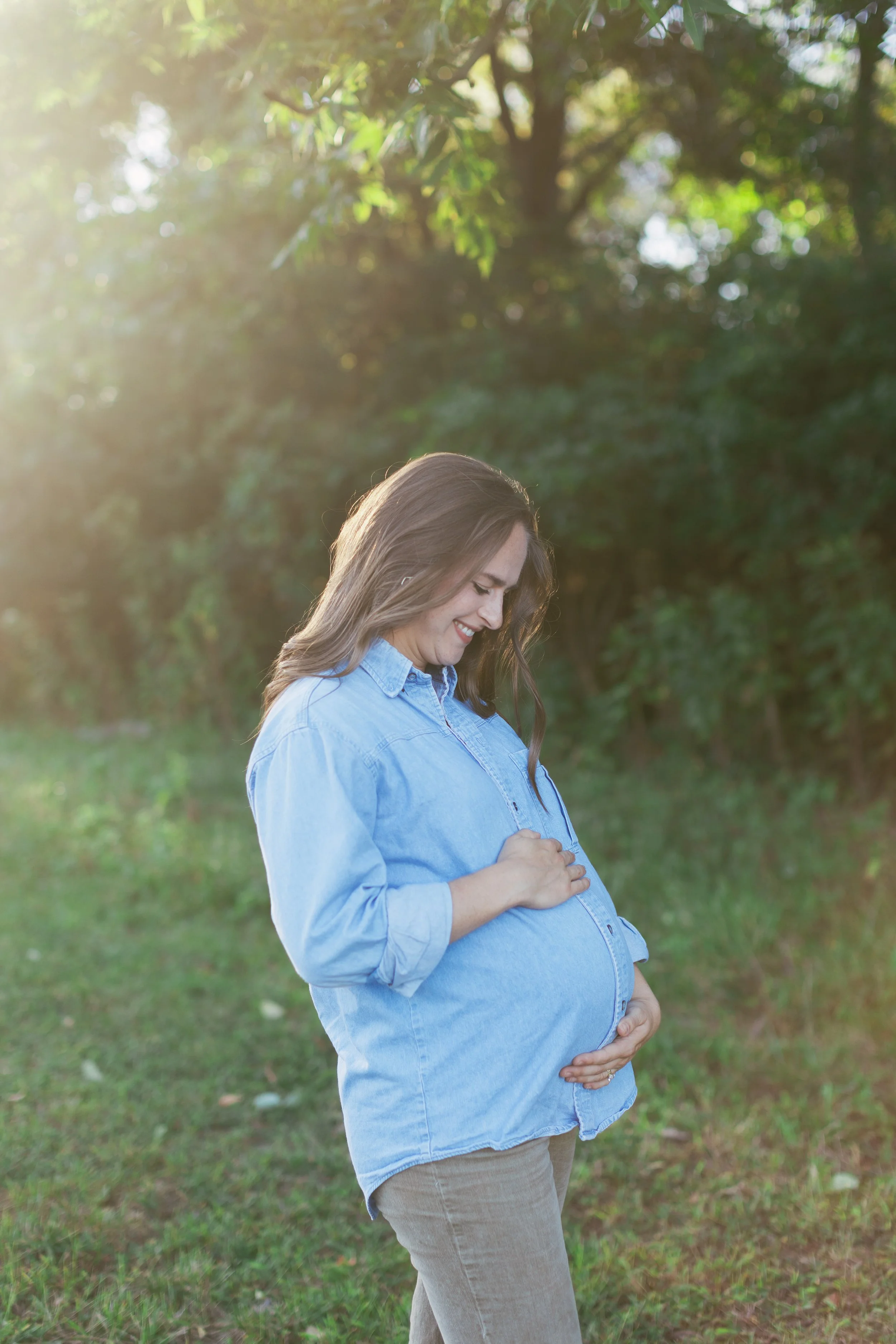 Pregnant woman standing outdoors in a green park, smiling and looking down at her baby bump with sunlight filtering through trees.