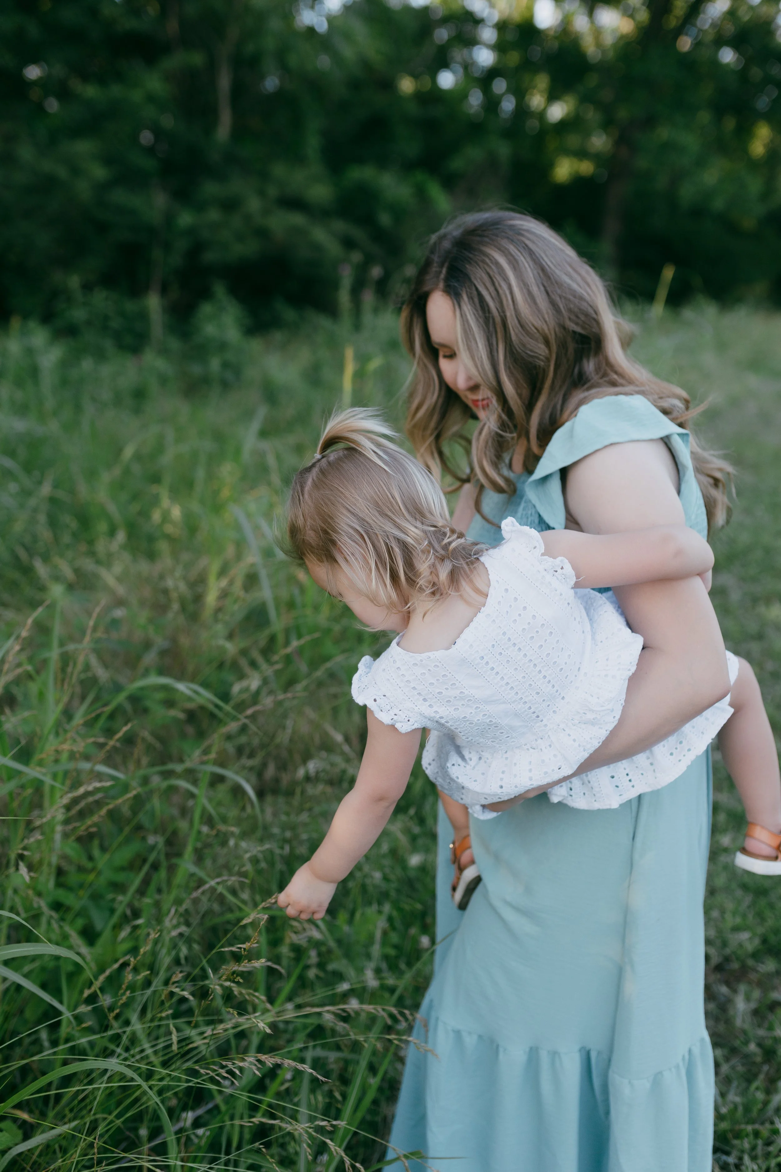 A woman holding a young girl in a white dress, with the girl reaching toward the grass in a lush outdoor setting.