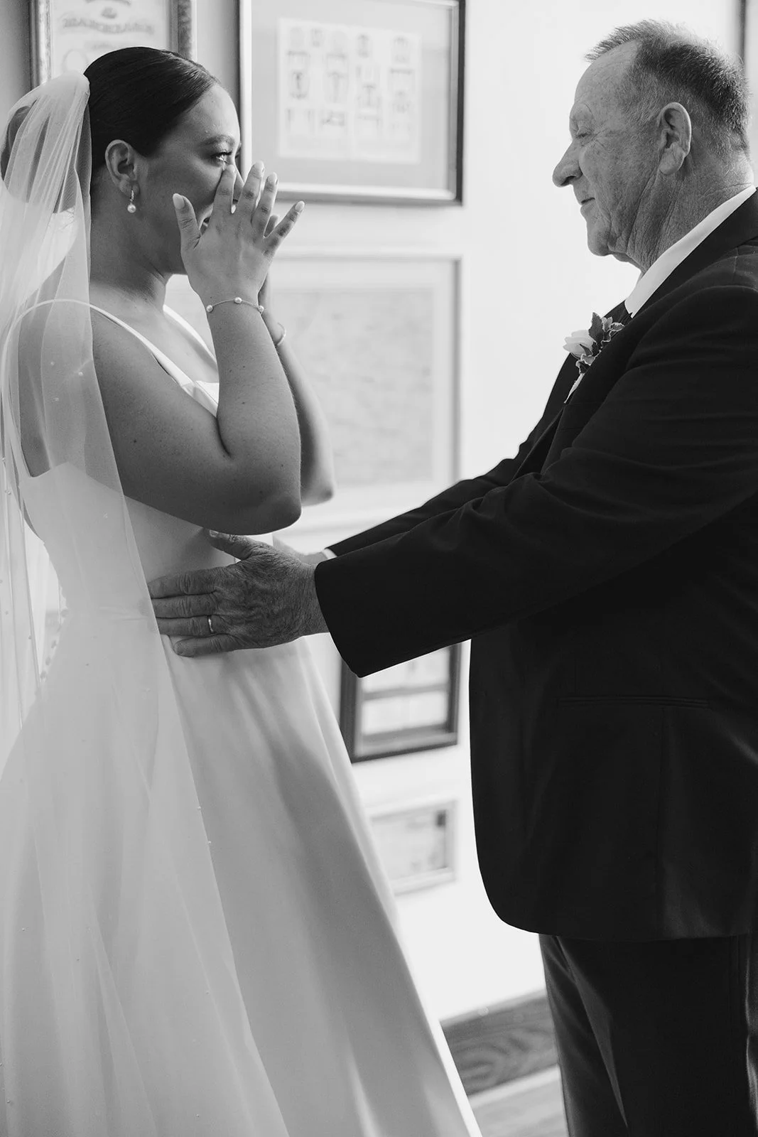 A bride in a wedding dress and veil is wiping tears as she is sharing a tender moment with her dad before her wedding ceremony.