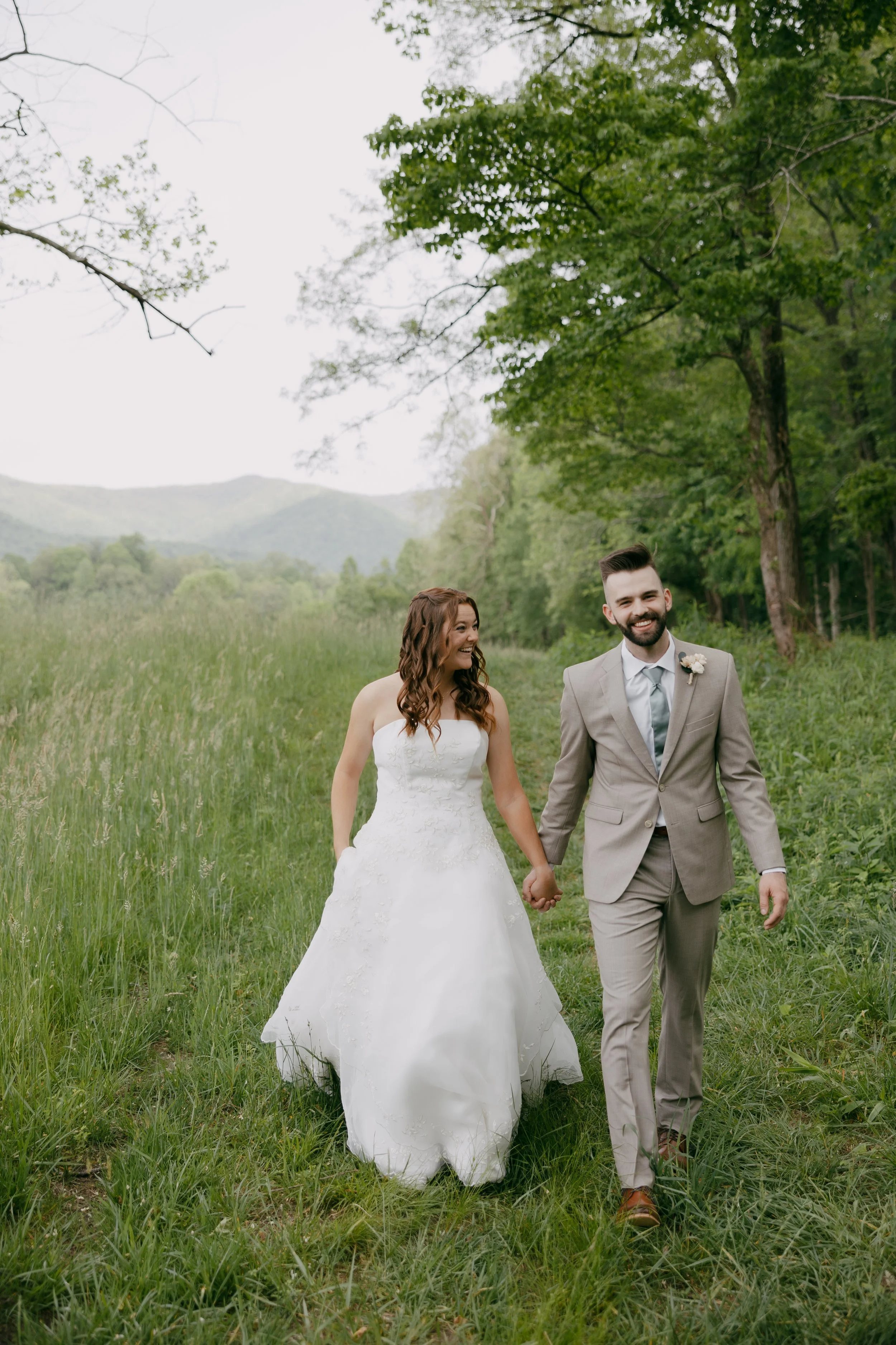 A bride and groom walking hand-in-hand through a grassy field surrounded by trees and mountains, smiling and enjoying their wedding day.