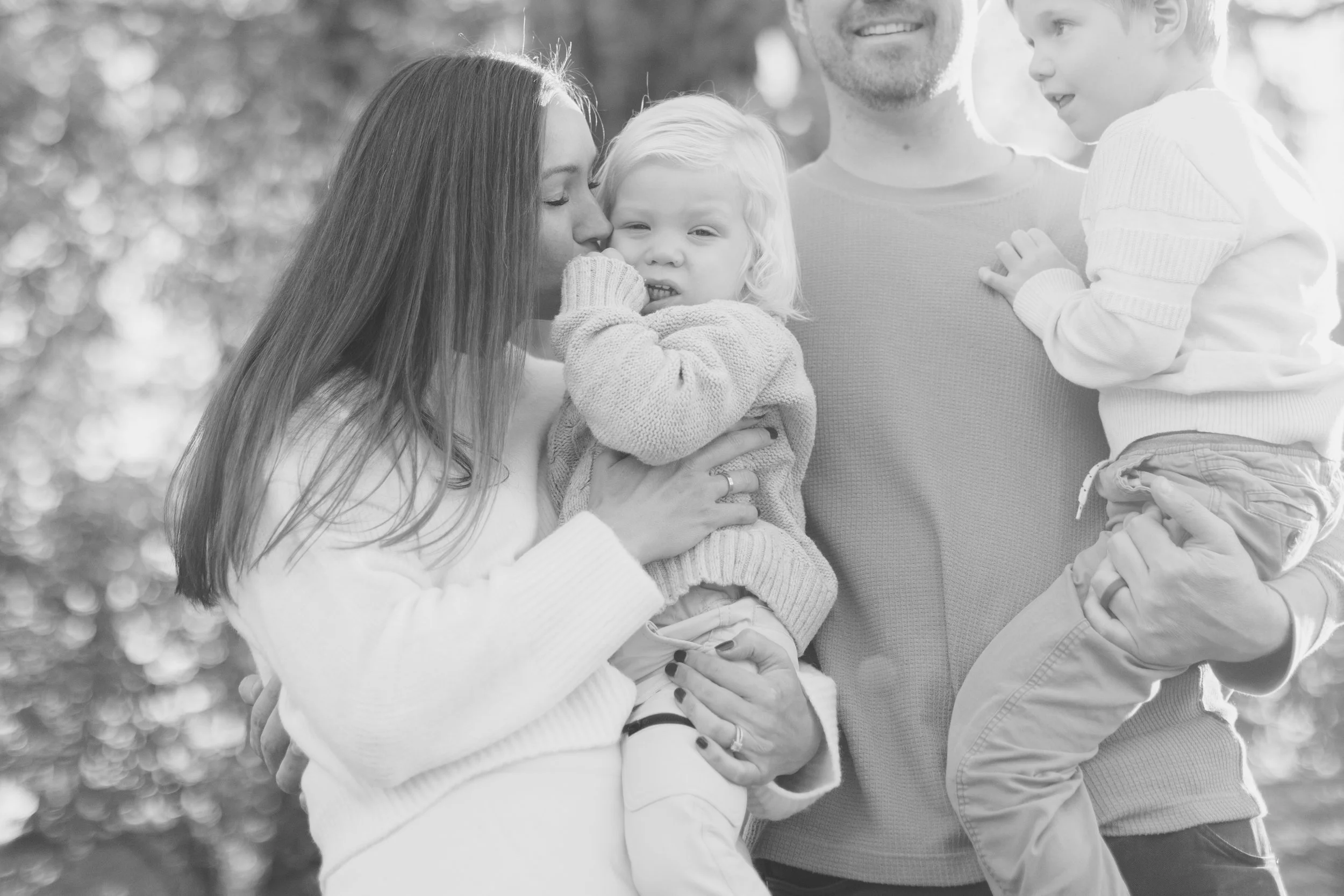A black and white photo of a happy family outdoors, with a woman holding a young child, a man holding another child, all smiling and close together.