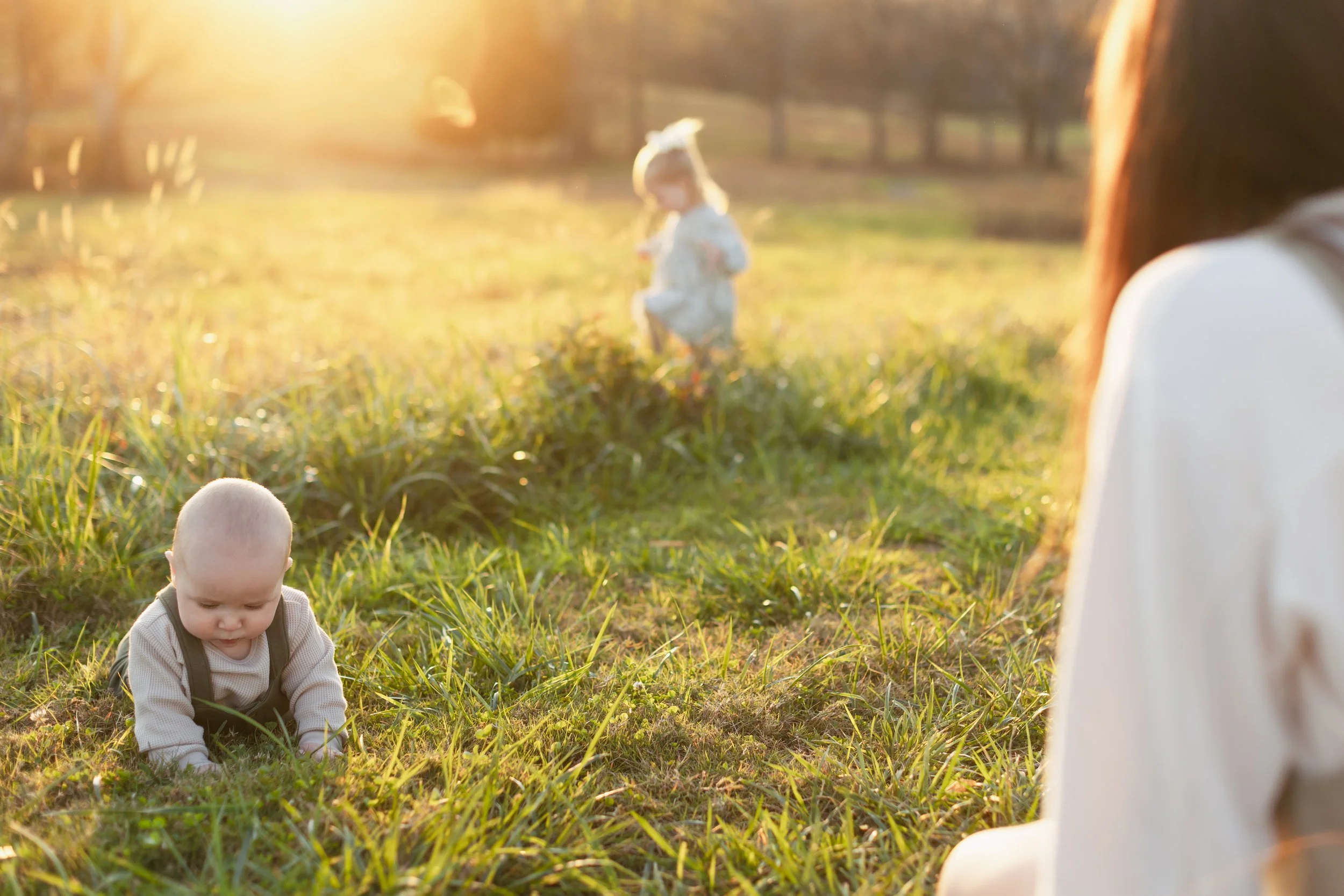 A woman sitting in a grassy field during sunset, watching her two young children playing. One child is crawling on the ground, and a girl is in the background running. The scene has warm golden lighting and a peaceful outdoor setting.