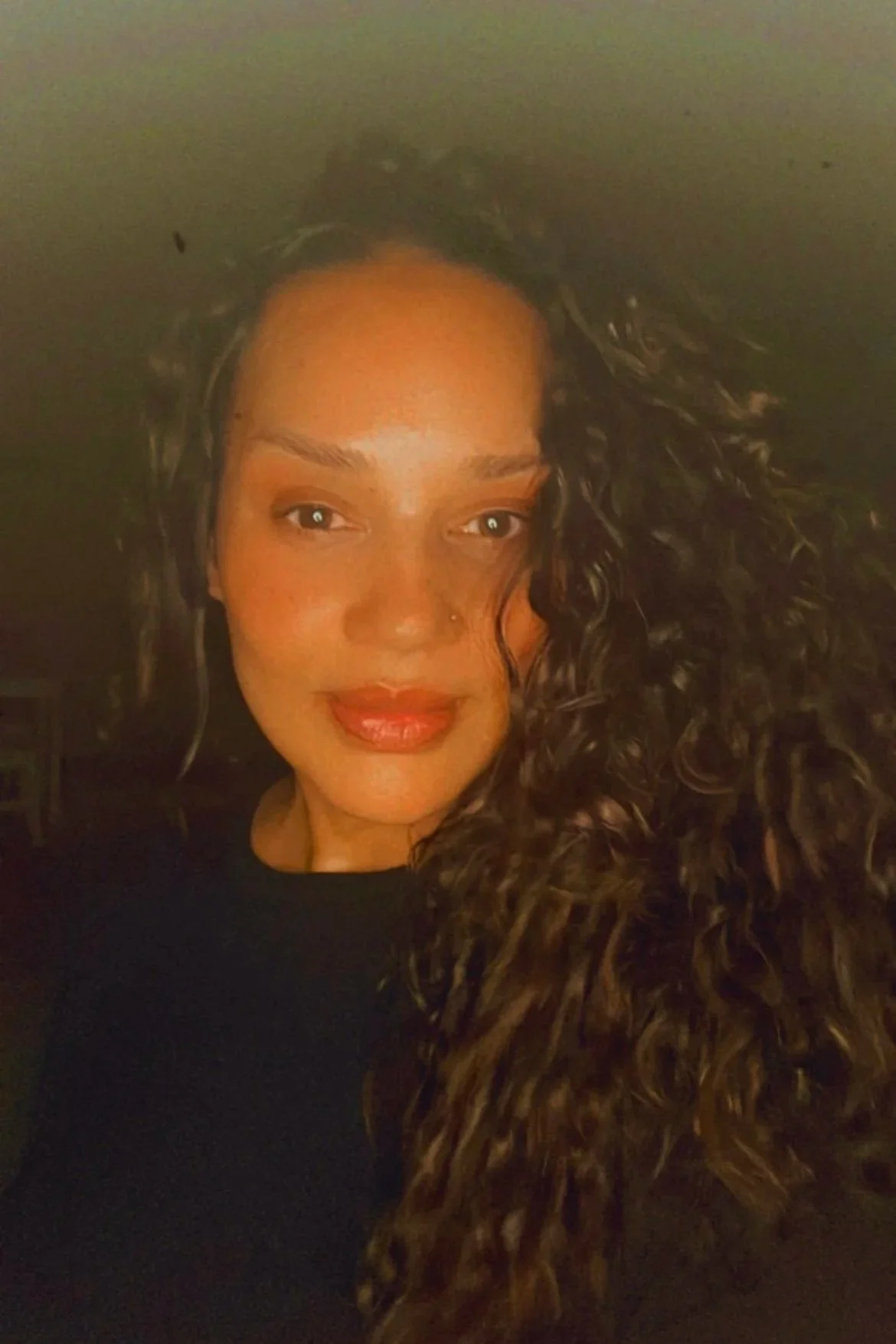 A woman with curly dark hair takes a selfie in a dimly lit setting. She has a nose piercing, light makeup, and is wearing a dark top.