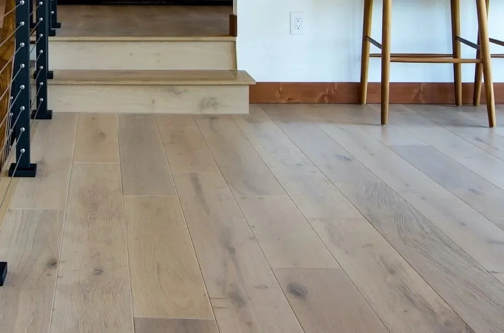 Interior view of a modern home with light wooden flooring, a small step up with two wooden steps, a black metal staircase with wooden steps, a high wooden table with bar stools, and a partial view of a gray sofa in the background.