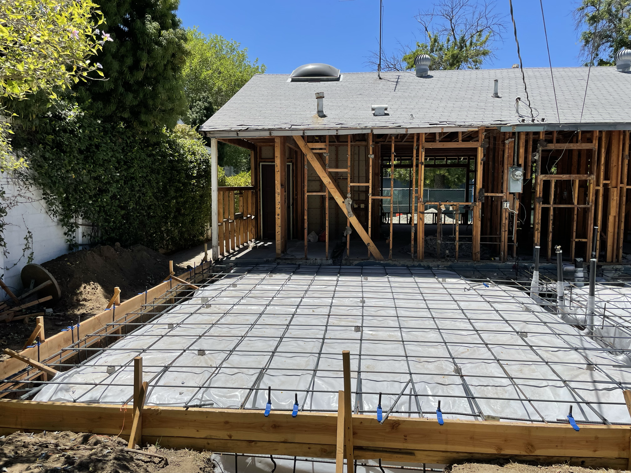 Construction site showing a house being renovated with a new concrete slab poured for a patio or foundation, construction rebar grid, and a partially framed wooden structure.