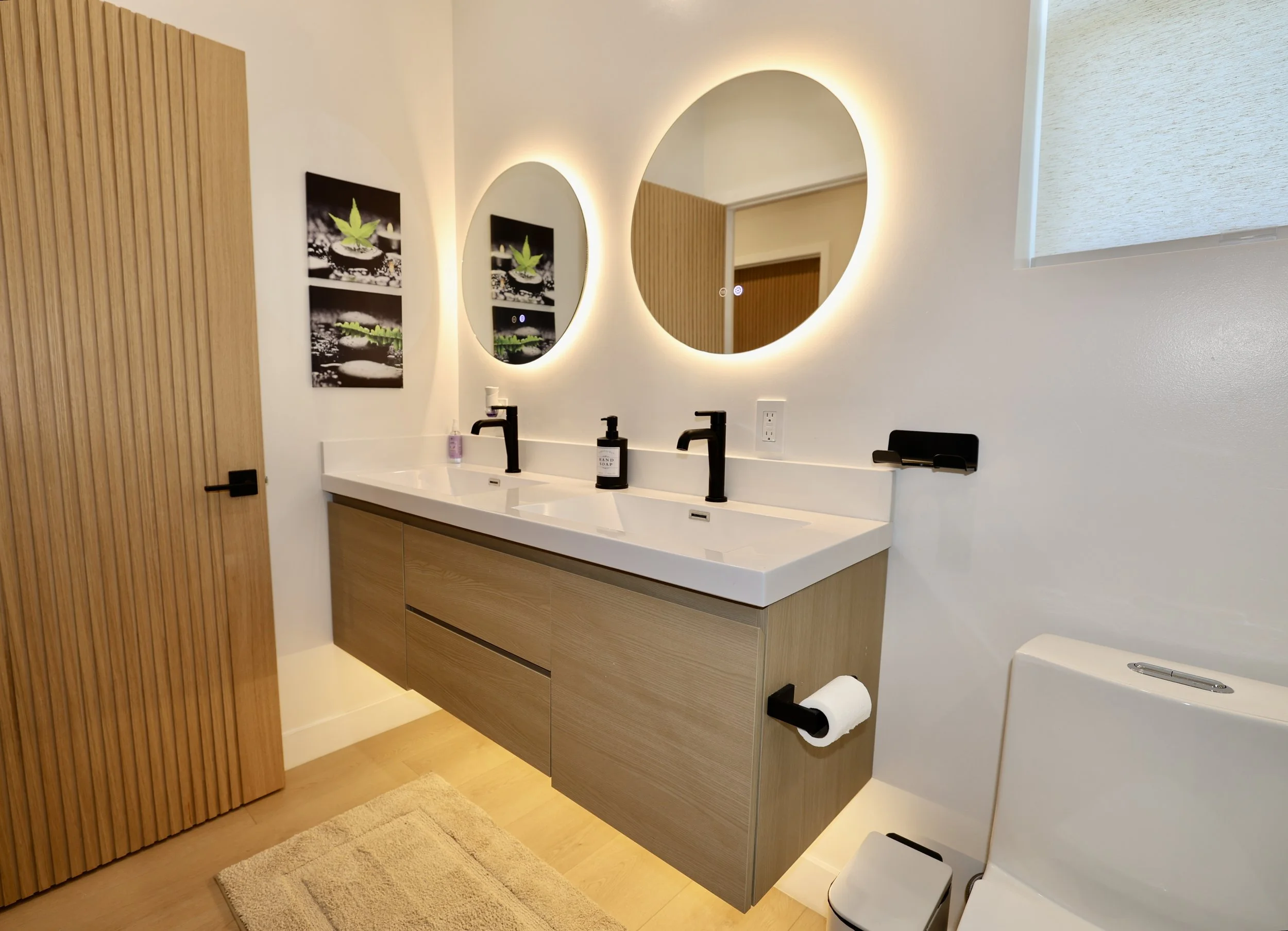 Modern bathroom with double sink vanity, round backlit mirrors, black faucets, and artwork of cannabis leaf on wall.