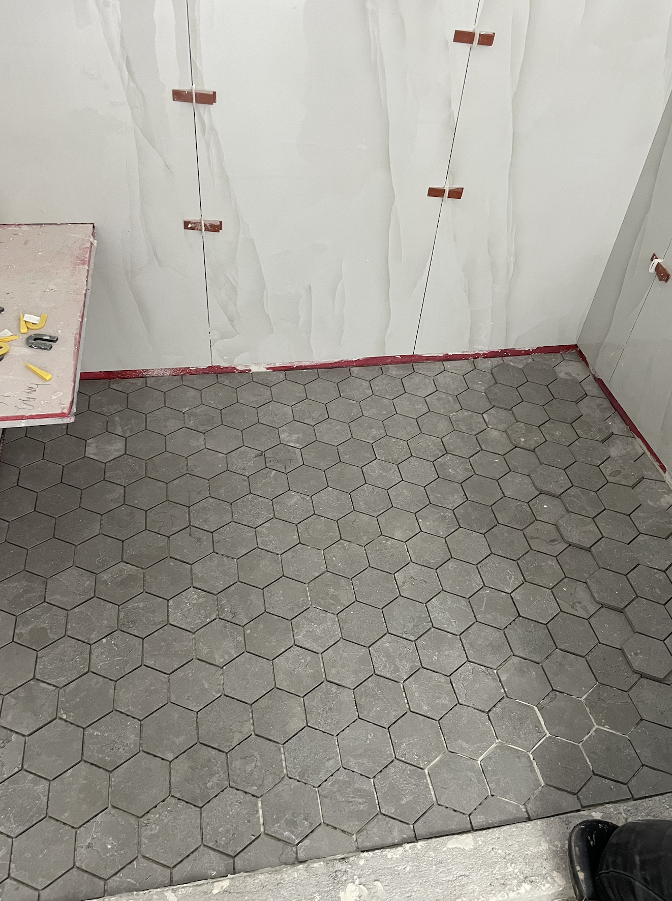 Hexagonal grey tiles on the floor of a room under construction with unfinished walls and red trim along the baseboards.