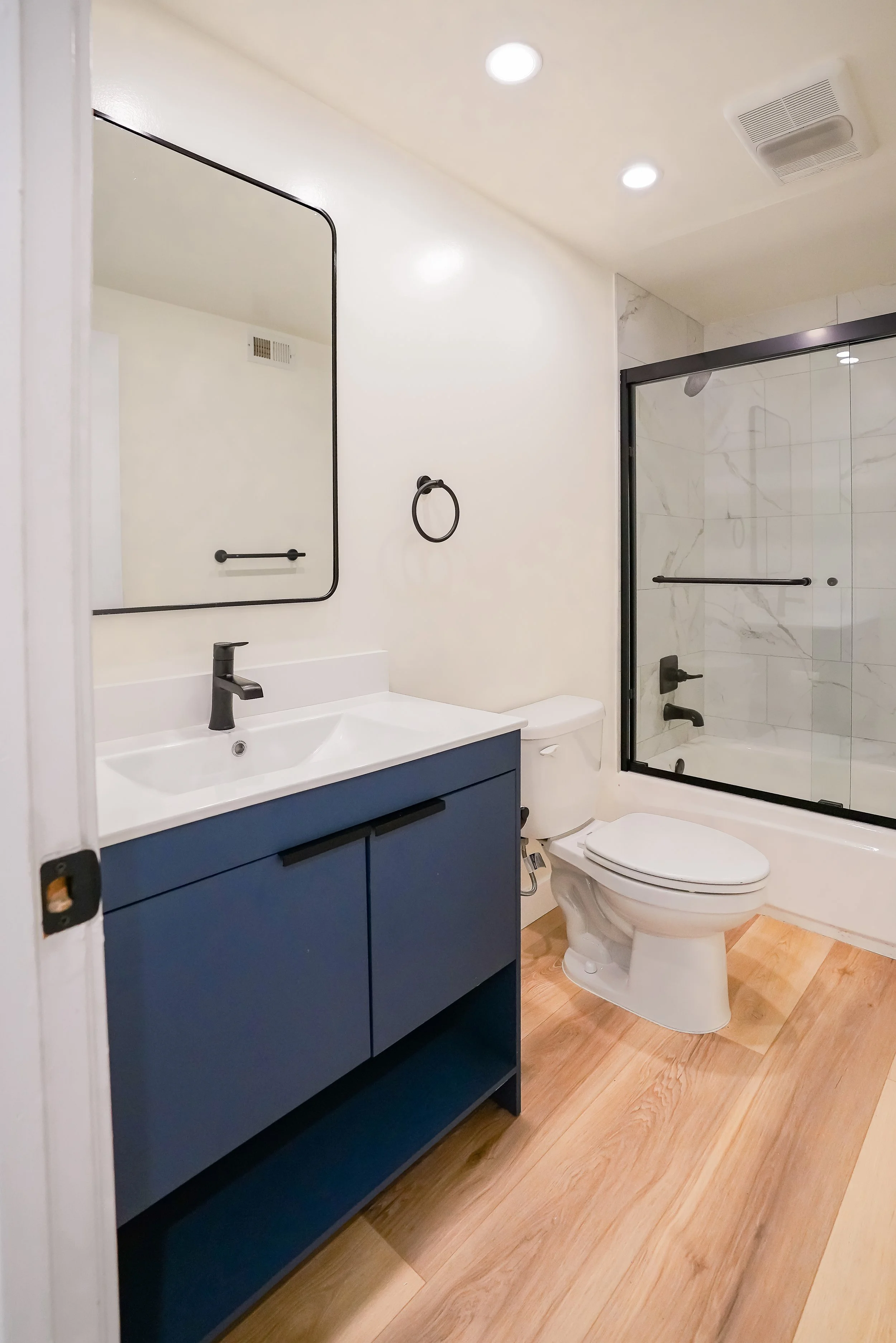 Modern bathroom featuring a blue vanity with a white sink, a round mirror, black fixtures, a white toilet, and a glass-enclosed shower with marble tiles.