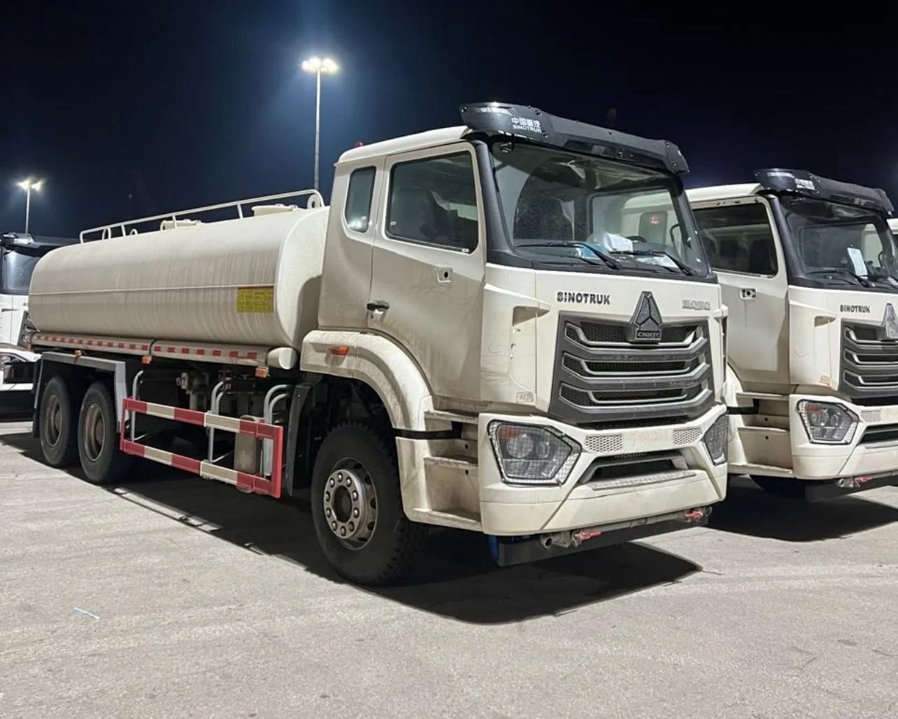 White Sinotruk tanker truck parked in a lot at night, with other trucks nearby and bright overhead lights illuminating the scene.