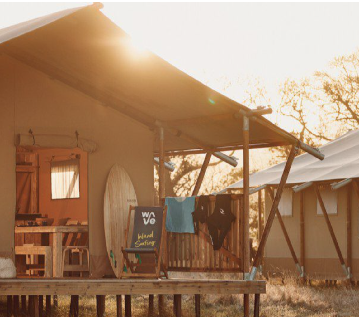 Sunlit outdoor deck area with a surfboard, a chalkboard sign reading 'wave', hanging clothes including a blue towel and black swimsuit, and a wooden dining area under a roof in a campsite setting with tents and trees in the background.