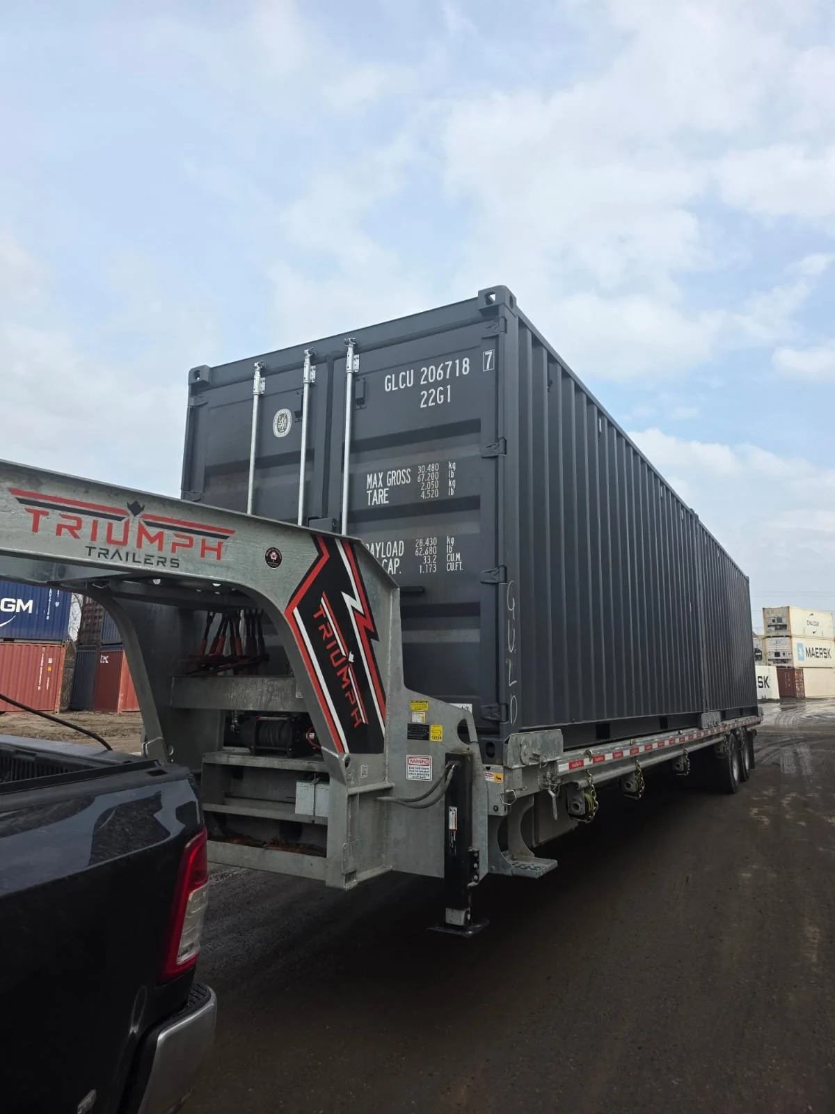 A large shipping container on a flatbed trailer being transported, with a Truck World tri-axle trailer hitch visible in the foreground, and additional stacked shipping containers in the background under a partly cloudy sky.