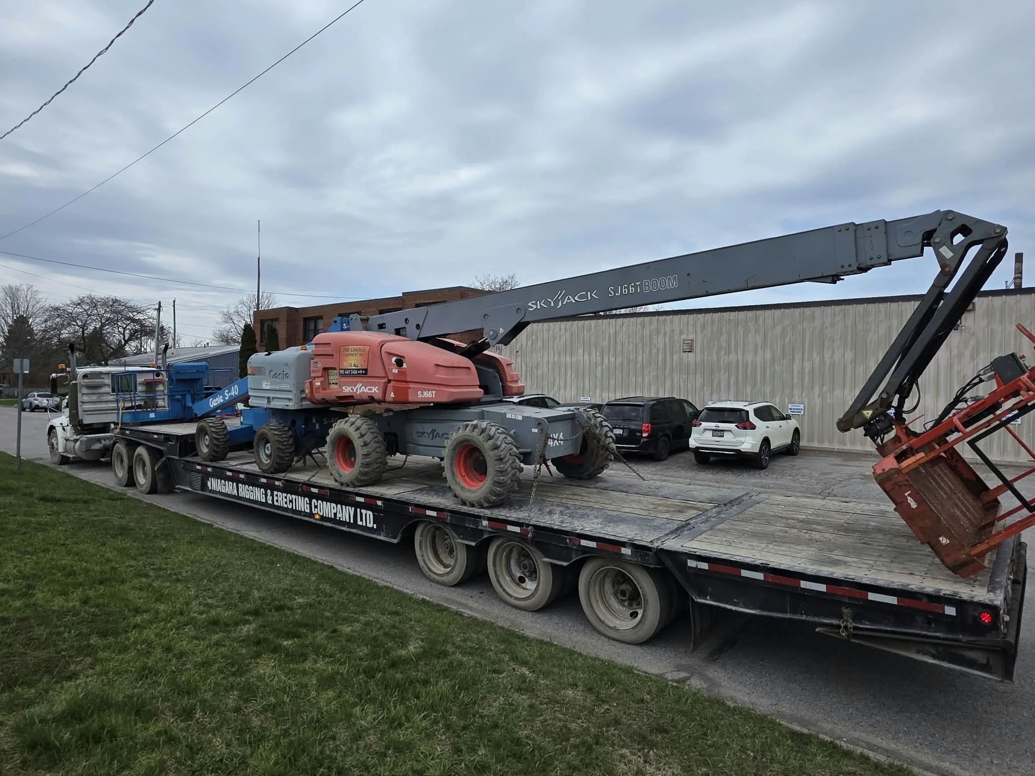 Flatbed truck transporting heavy construction equipment, including a large red and gray telescopic boom lift, labeled Skyjack, and a blue Genie lift, parked in front of a brick building under cloudy sky.