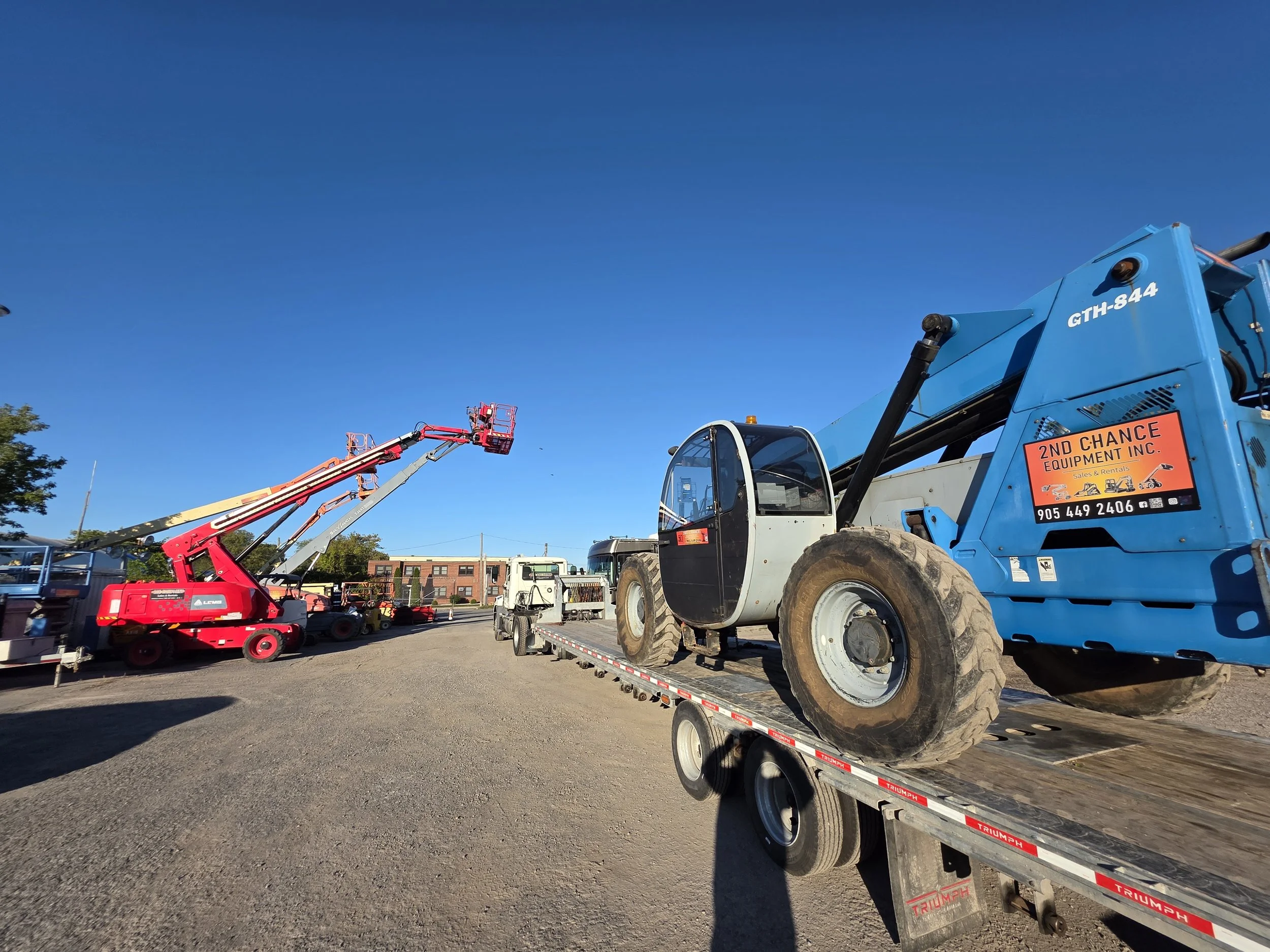 Construction equipment including a blue crane on a flatbed truck carrying a compact excavator and other machinery in a lot, under a clear blue sky.