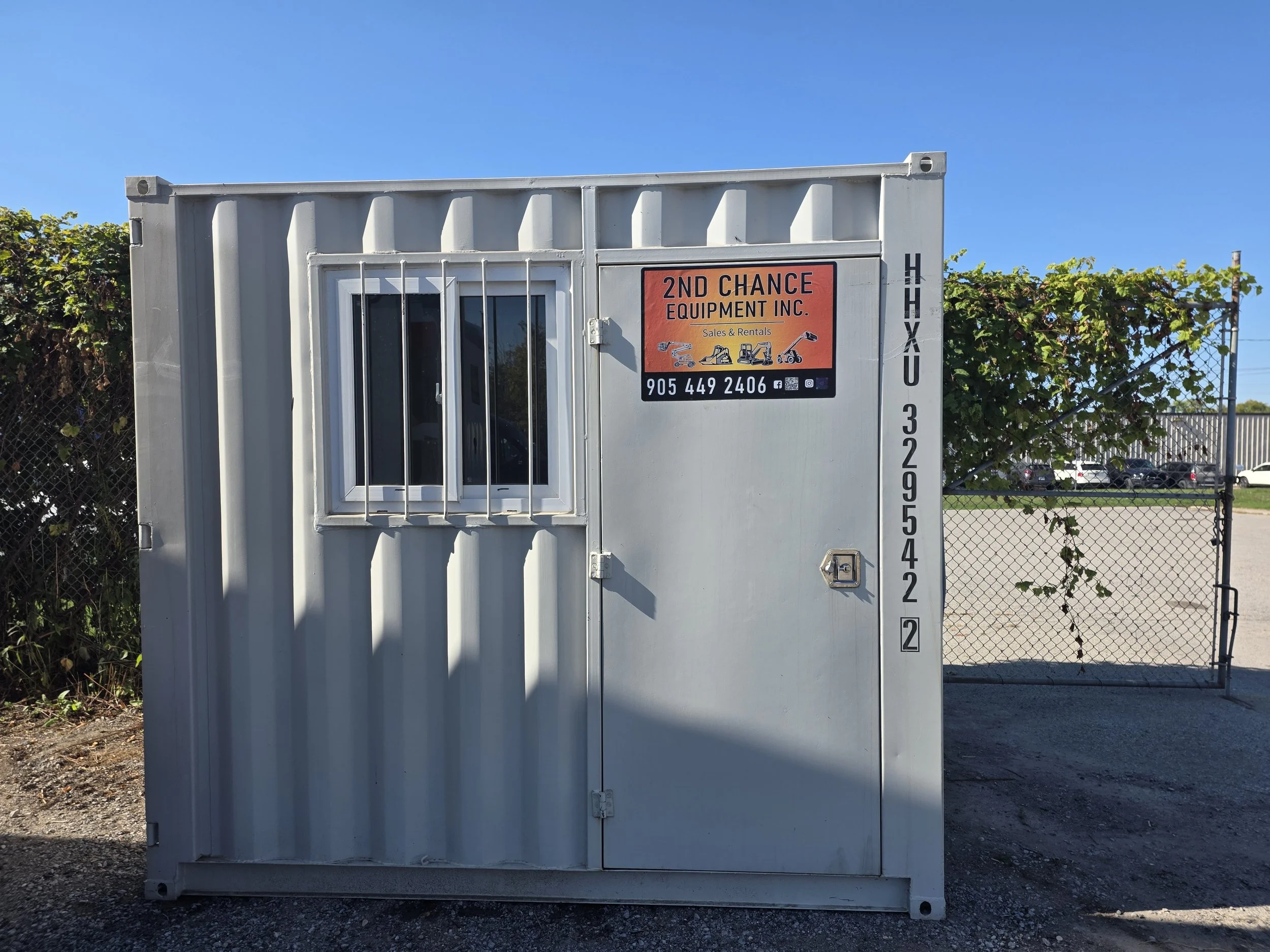 A white shipping container with a small window secured by metal bars. There is a sign on the container that reads "2nd Chance Equipment Inc. Sales & Rentals" with icons of equipment and a phone number. The container is outdoors, with a chain-link fence and some greenery in the background under a clear blue sky.