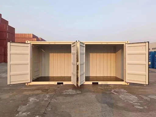 Two empty beige shipping containers with open doors, standing on a paved surface outdoors.