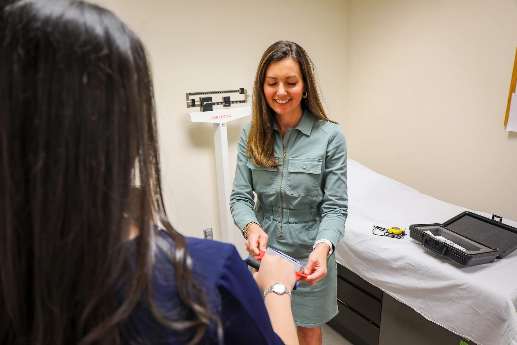 Woman in gray medical coat smiling and handing a blood pressure cuff to another woman in a hospital room with a bed and medical equipment.