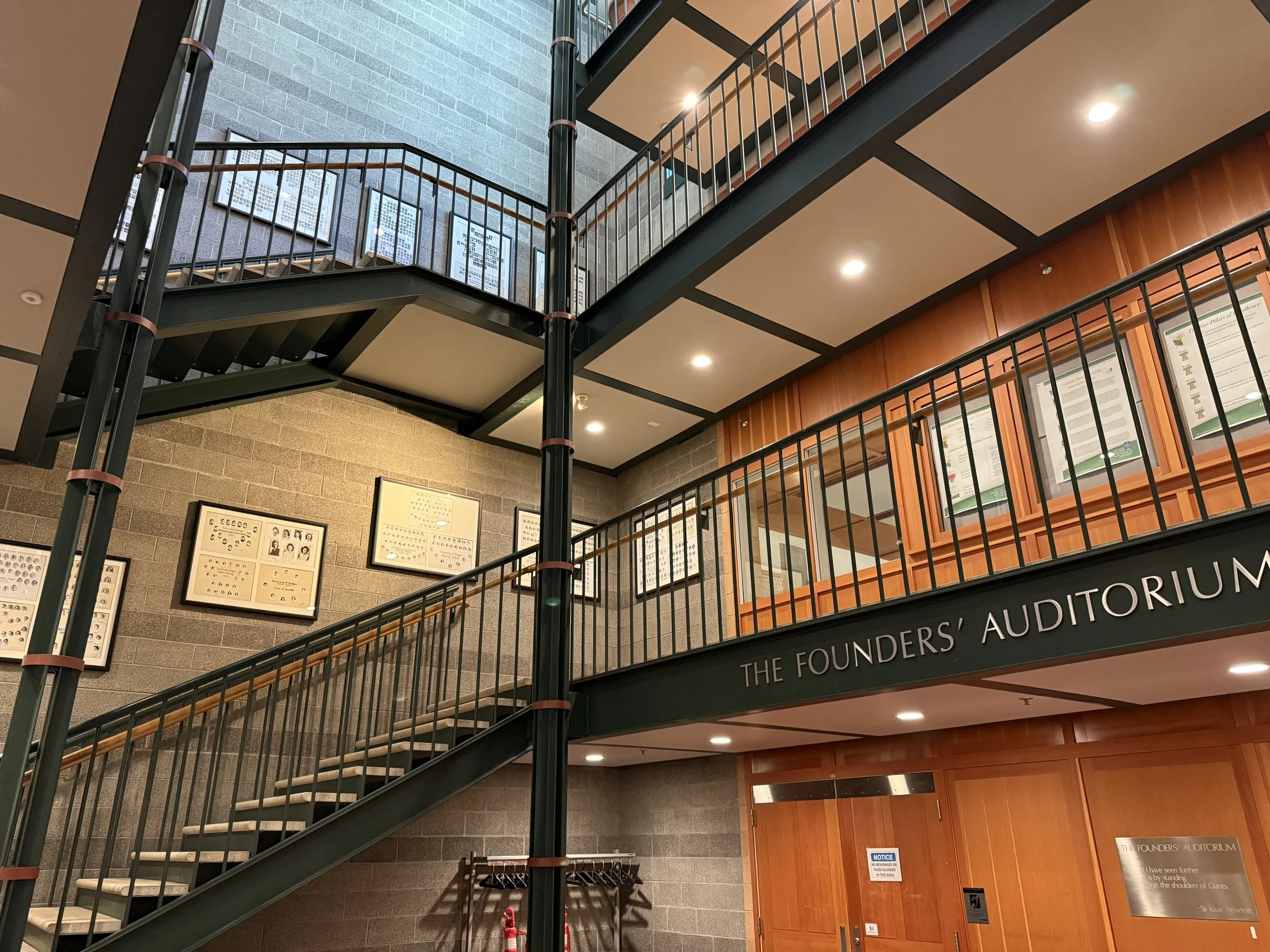 Interior view of a multi-level atrium in a building called 'The Founders' Auditorium,' featuring staircases, framed pictures on brick walls, informational posters, and wooden wall panels.