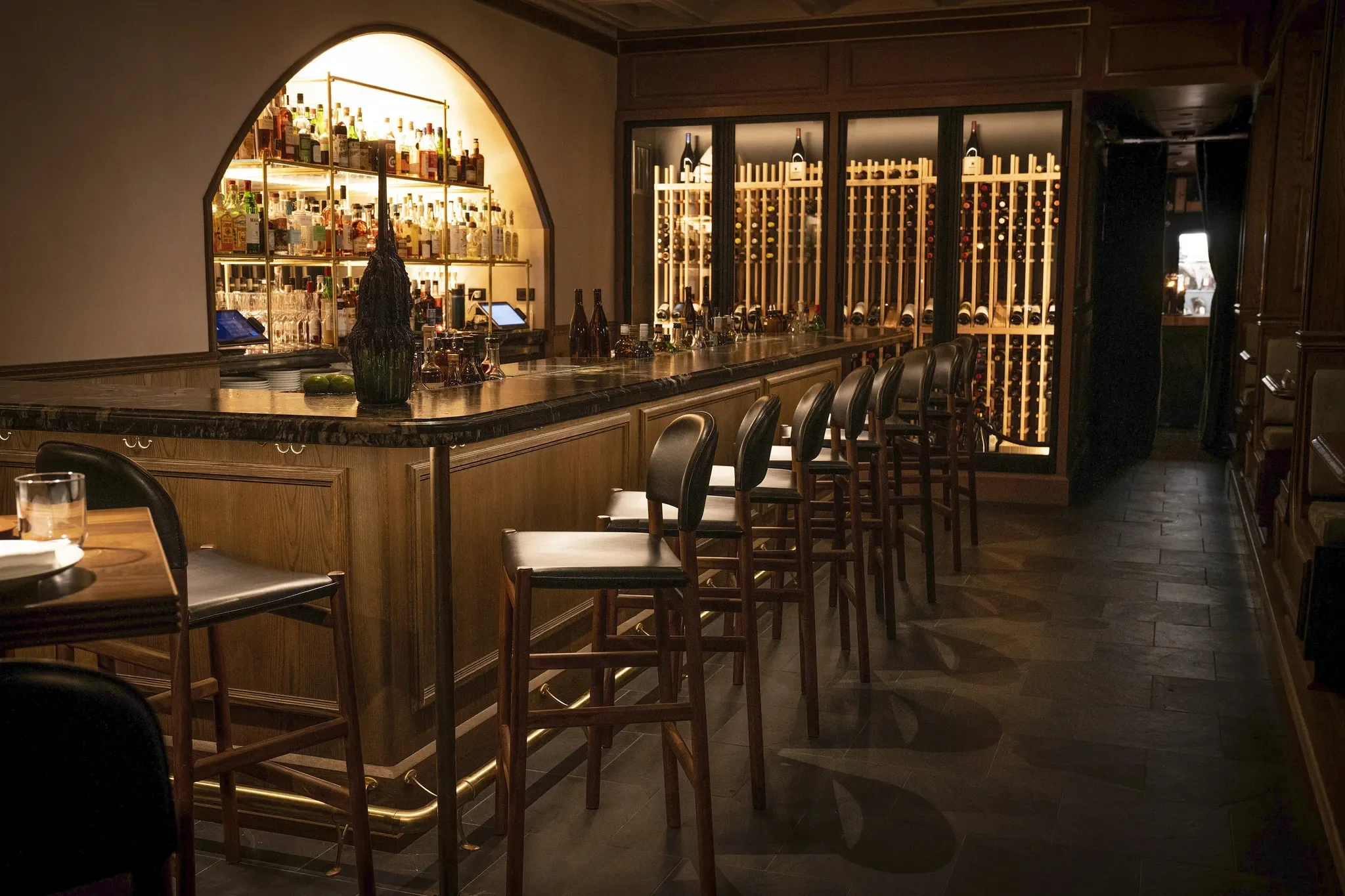 Elegant bar with shelves filled with bottles, a long wooden counter, and leather bar stools in a cozy, warmly lit setting.