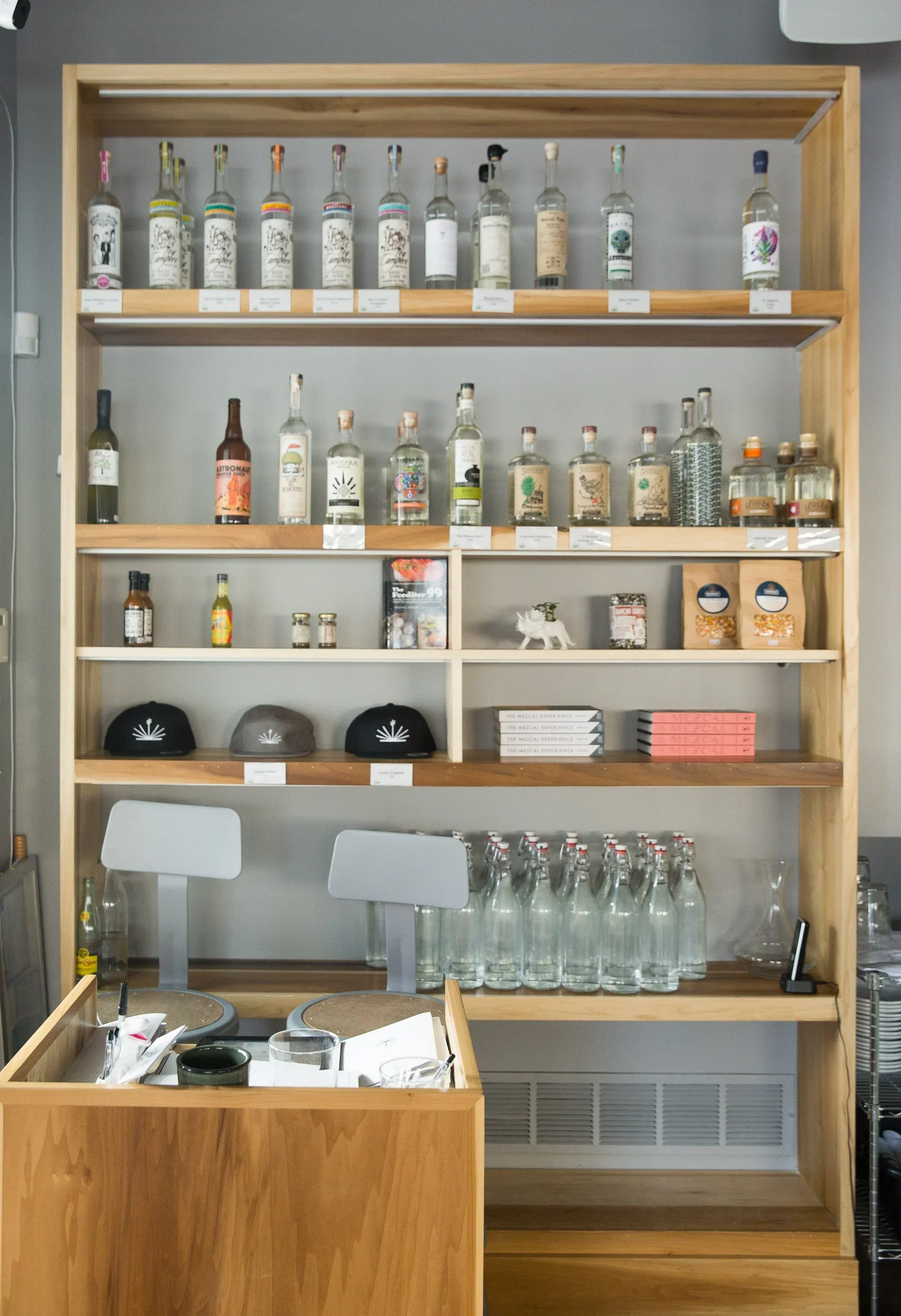 Interior of a bar with a white counter, black chairs, and a shelf with bottled drinks. A person is sitting at the bar. Artwork is on the wall.