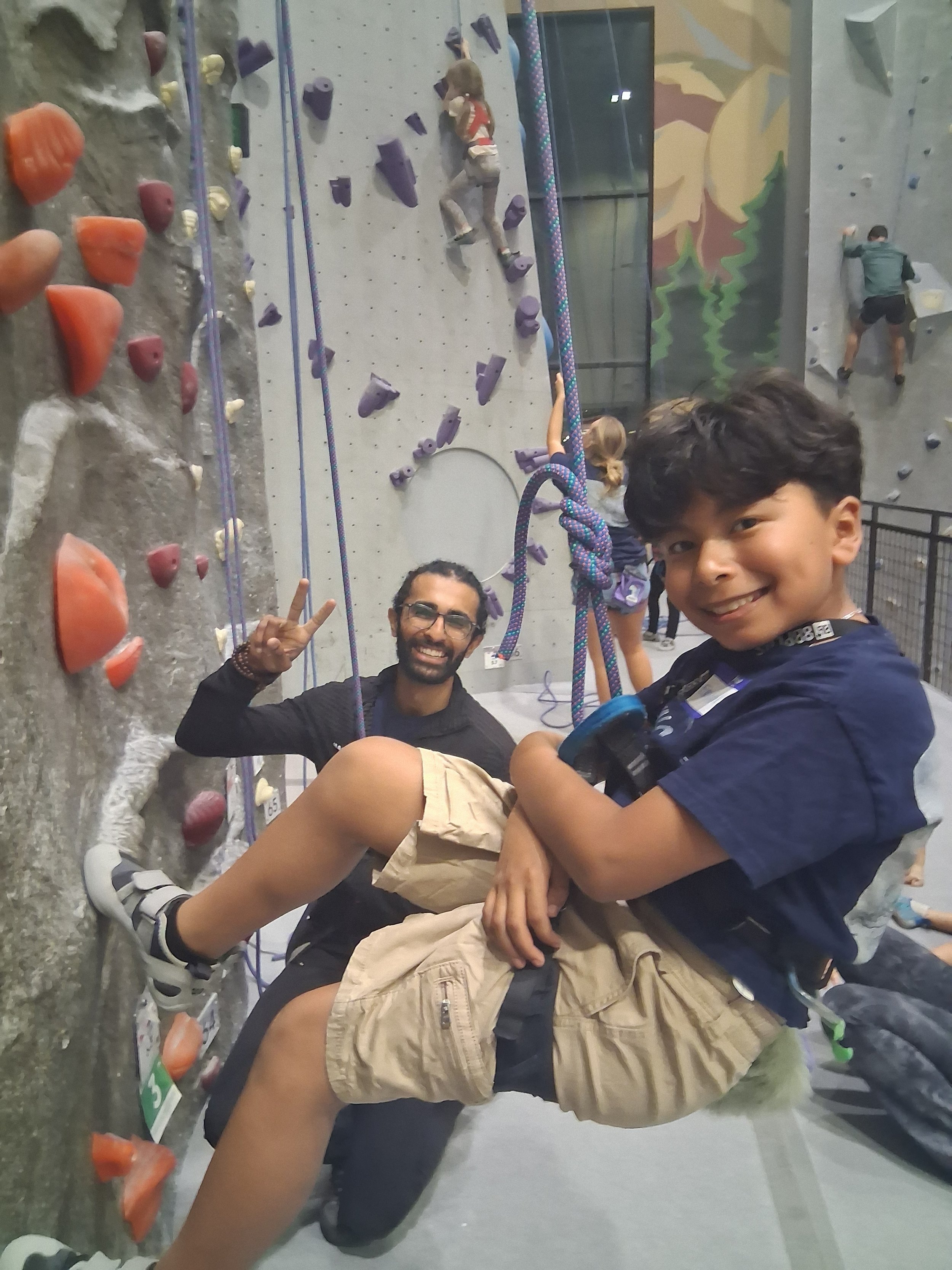 A smiling young boy with dark hair in a navy t-shirt and beige shorts sitting on a rock climbing wall, with an adult man crouching beside him making a peace sign, at an indoor rock climbing gym.