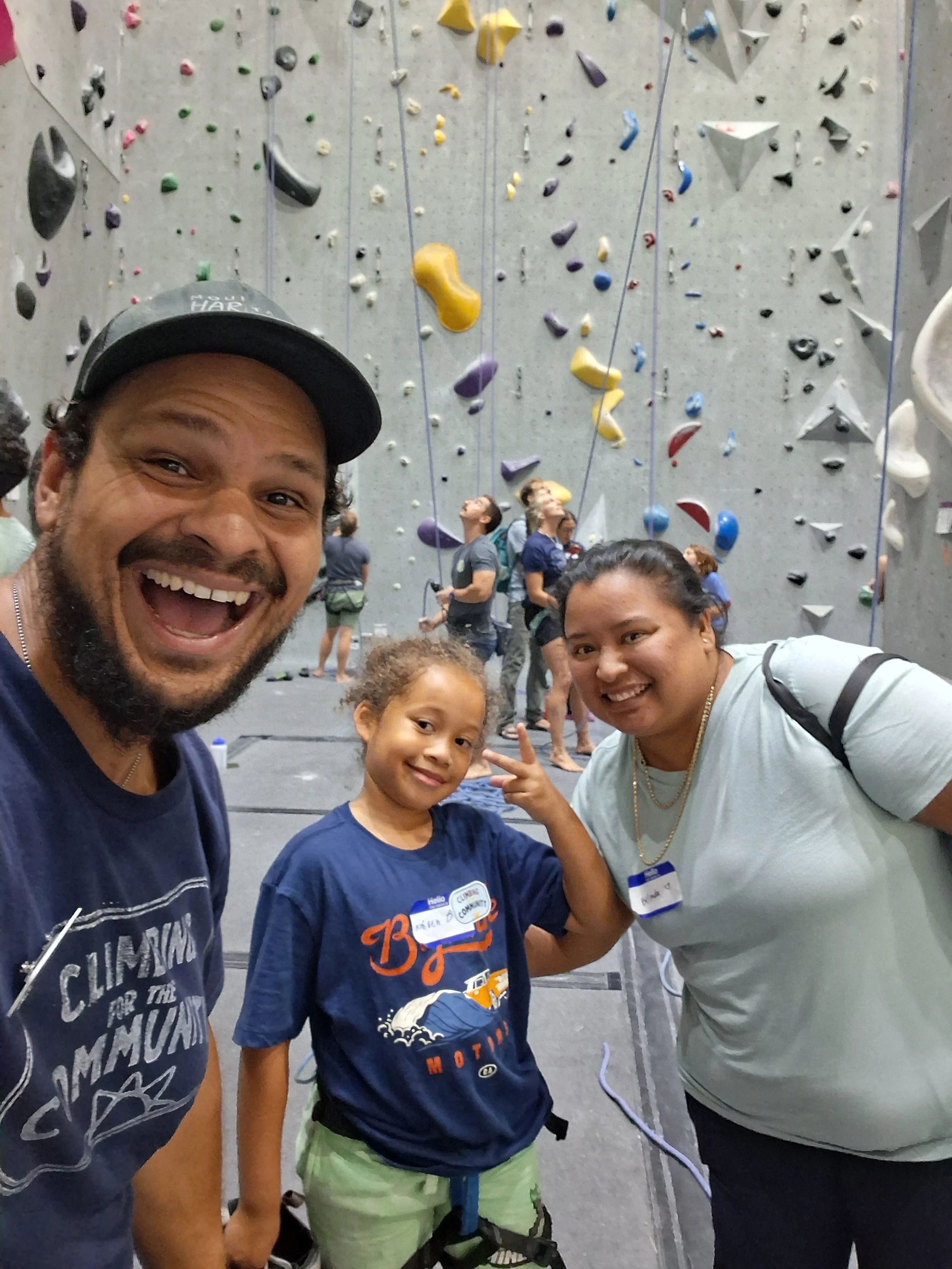 A group of three smiling people, two adults and a child, posing for a selfie at an indoor rock climbing gym, with a climbing wall and other climbers in the background.