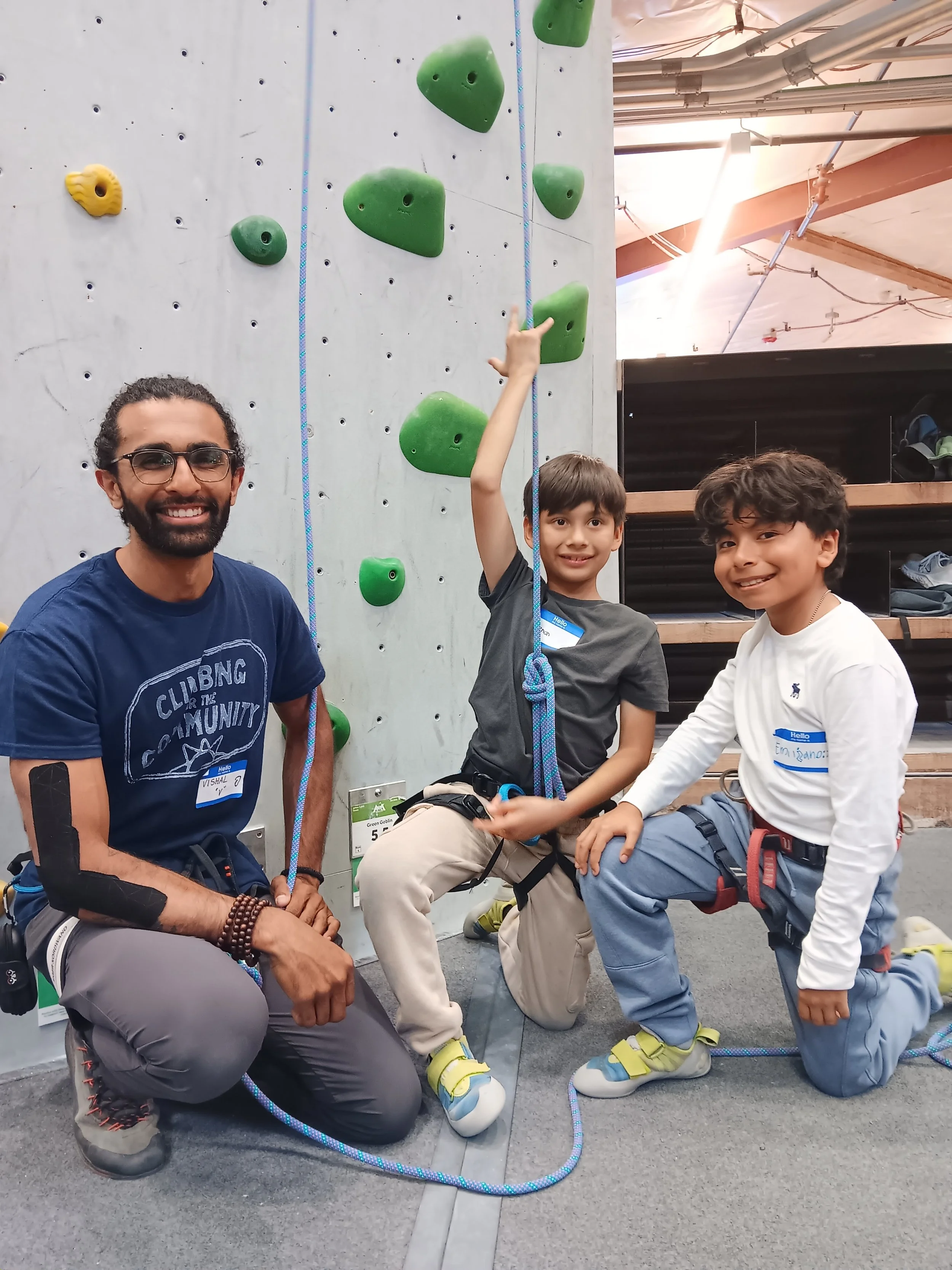 Two young boys and an adult male at an indoor rock climbing gym, kneeling in front of a climbing wall with green holds. The boy in the gray shirt is pointing upward, and the other boy in a white shirt is smiling. They are all wearing harnesses and cl