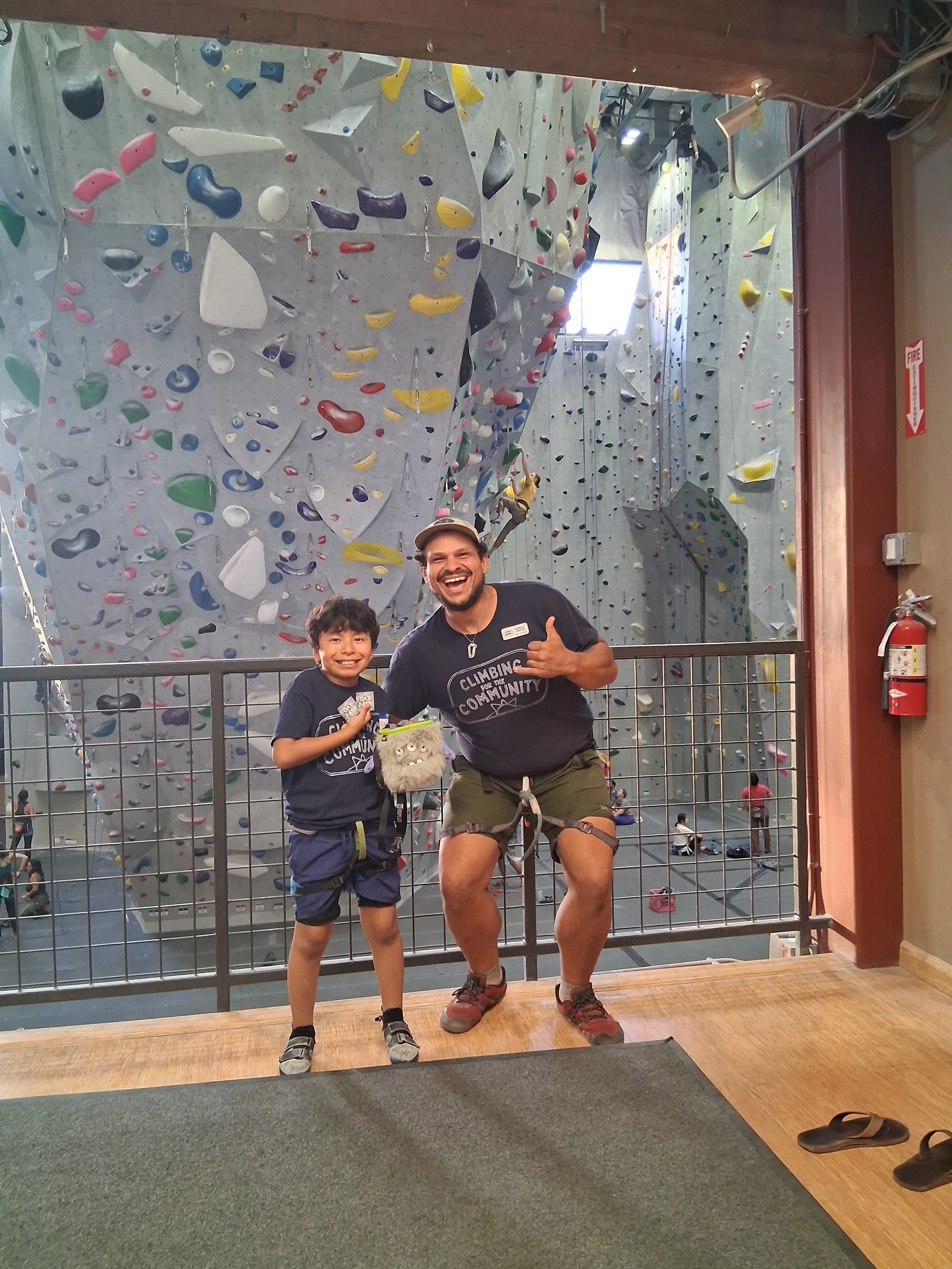 A man and a boy standing in front of an indoor climbing wall, smiling and posing for the camera. The boy is holding a small stuffed animal.